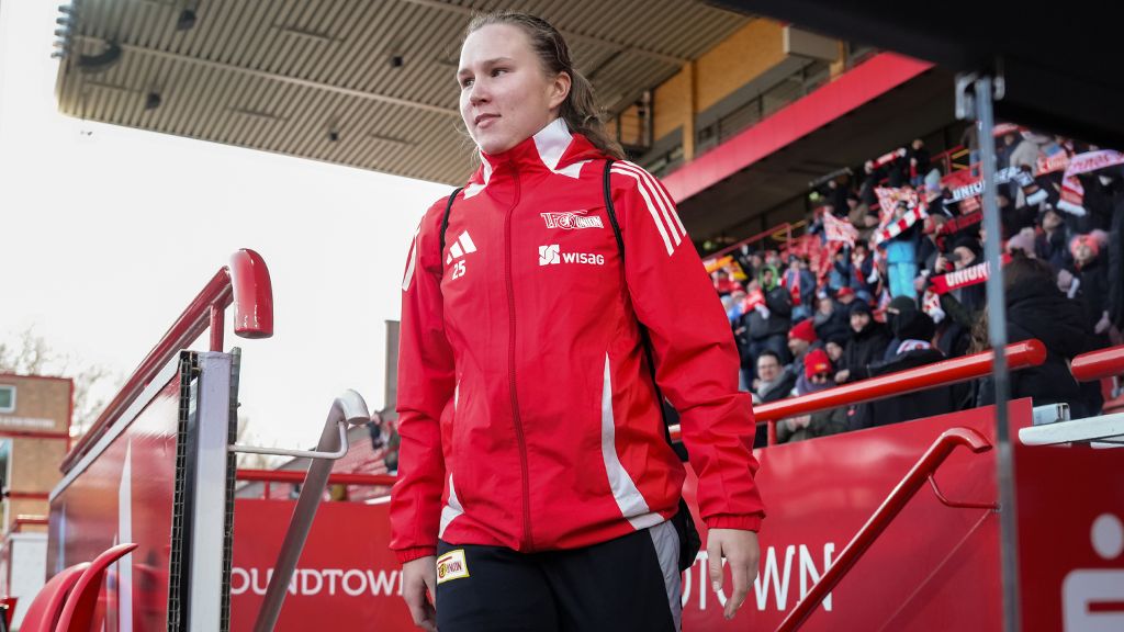 Eine Sportlerin in roter Jacke betritt ein Stadion, umgeben von jubelnden Fans im Hintergrund.