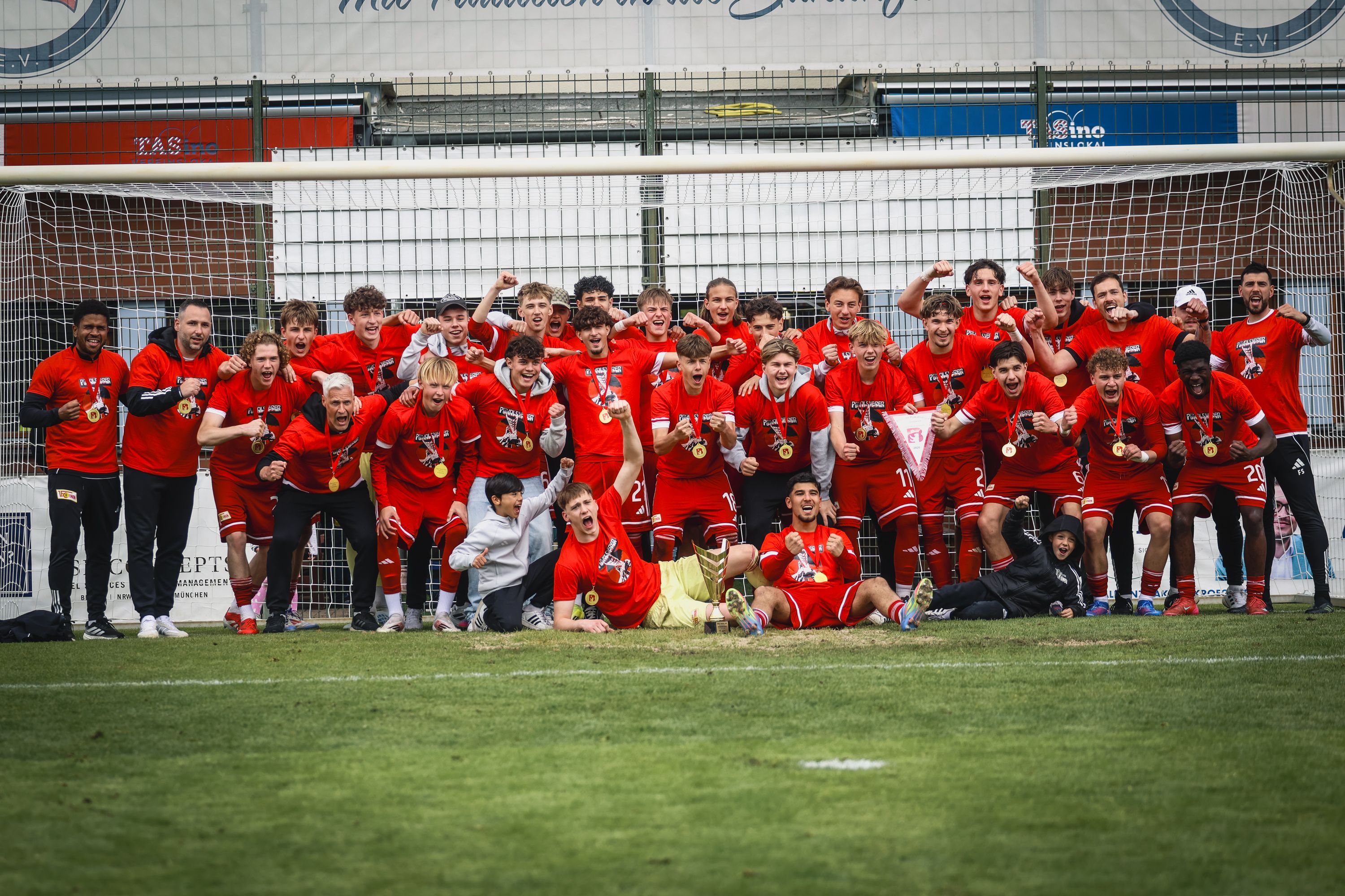 Eine Gruppe von Fußballspielern in roten Trikots feiert mit Medaillen vor einem Tor.