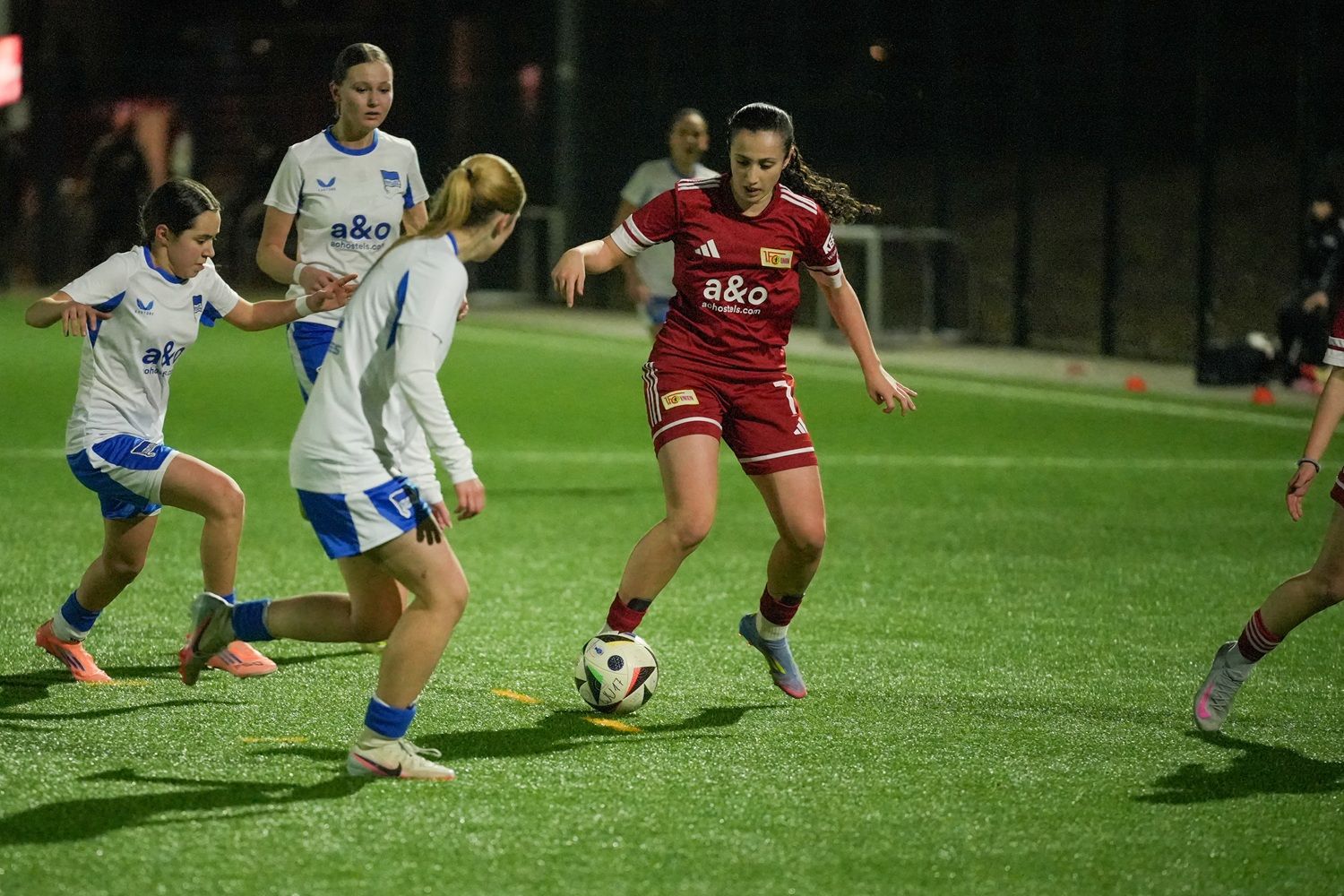 Eine Fußballspielerin in rotem Trikot dribbelt den Ball, während sie von zwei Gegnerinnen in weißen Trikots angegriffen wird.