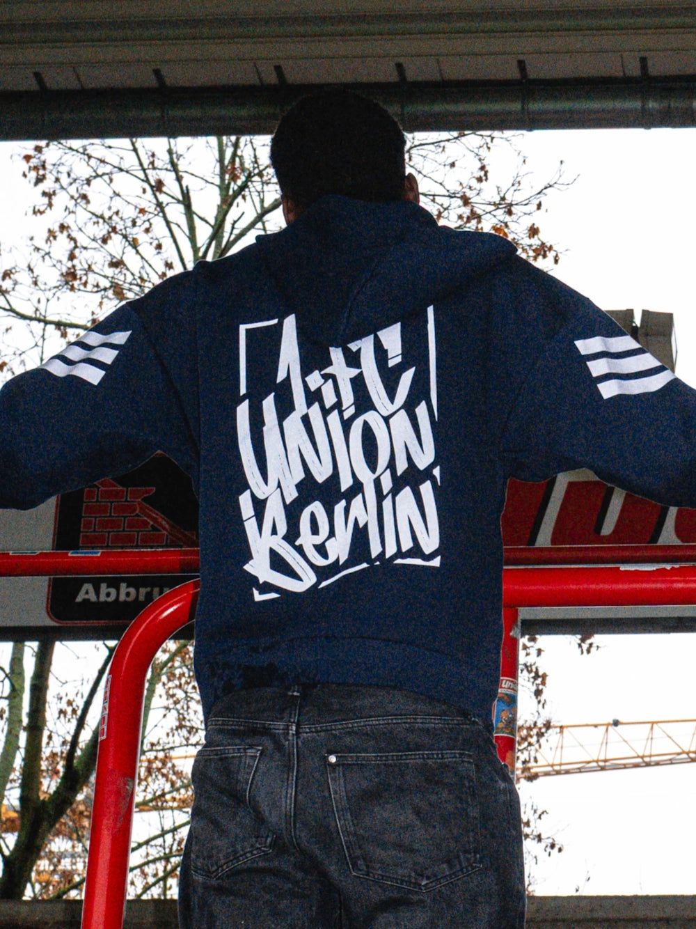 Person mit grauem Kapuzenpullover mit dem Aufdruck "Union Berlin" auf dem Rücken, steht vor einer roten Struktur.