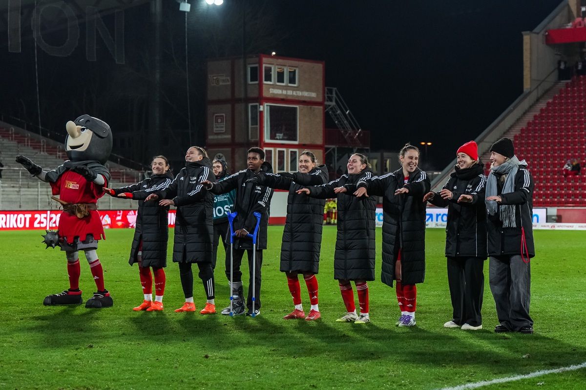 Eine Gruppe von Fußballspielerinnen und einem Maskottchen steht auf dem Spielfeld und applaudiert.