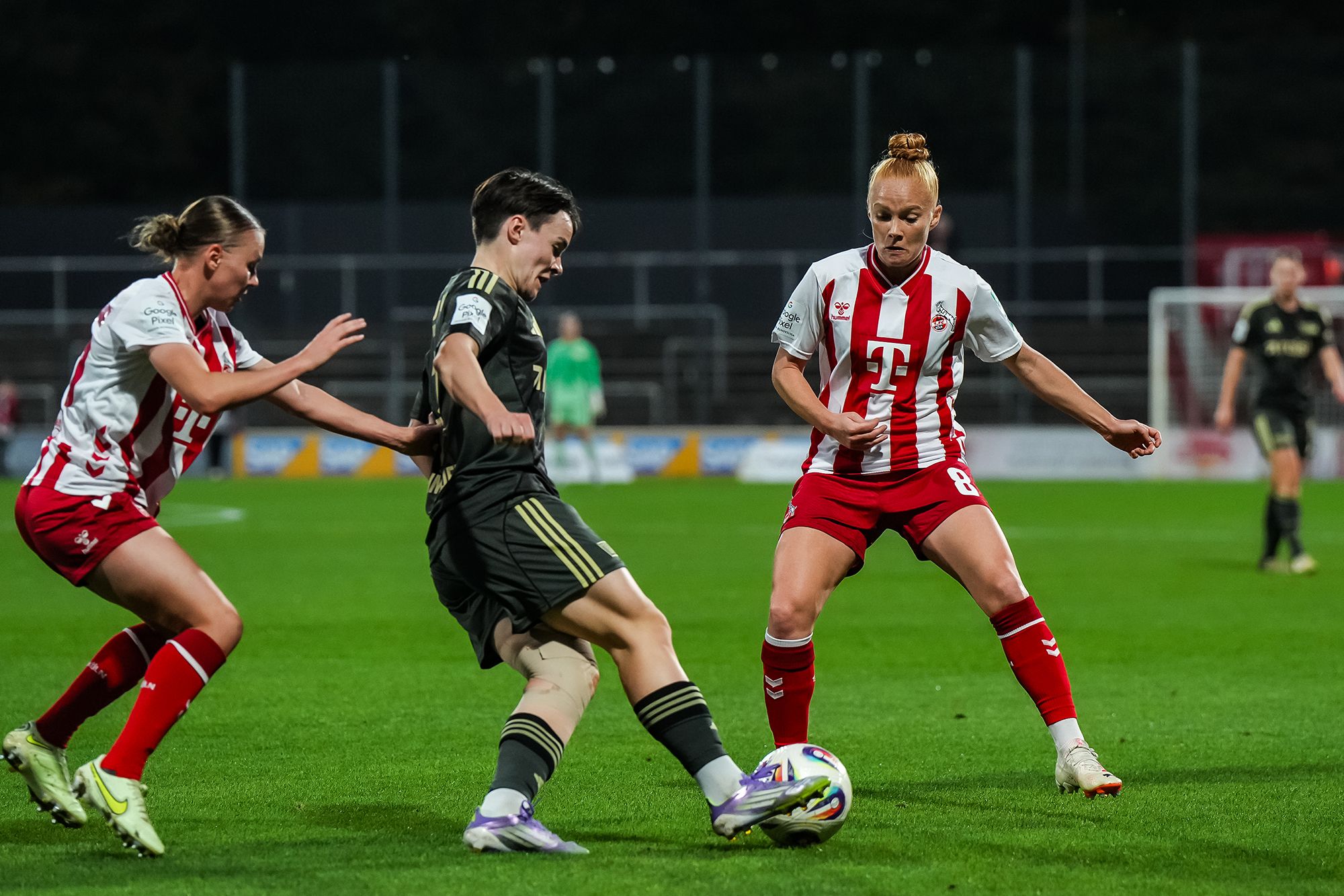 Zwei Spielerinnen in roten und weißen Trikots versuchen, den Ball von einer Spielerin in schwarzem Trikot zu erobern.