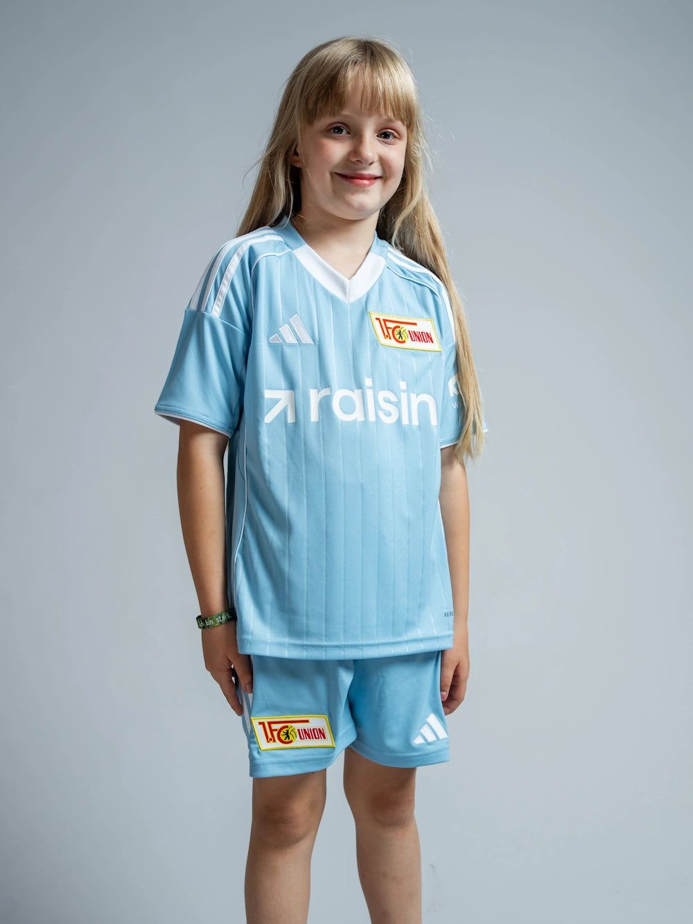 Ein Mädchen trägt ein hellblaues Fußballtrikot mit der Aufschrift "raisin" und passenden Shorts, mit einem freundlichen Lächeln.