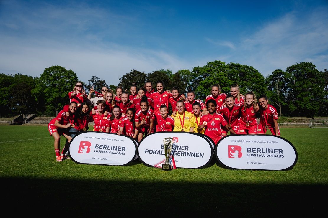Eine Gruppe von Frauen in roten Fußballtrikots pose für ein Gruppenfoto auf einem Sportplatz mit Pokalen und Bannern im Hintergrund.