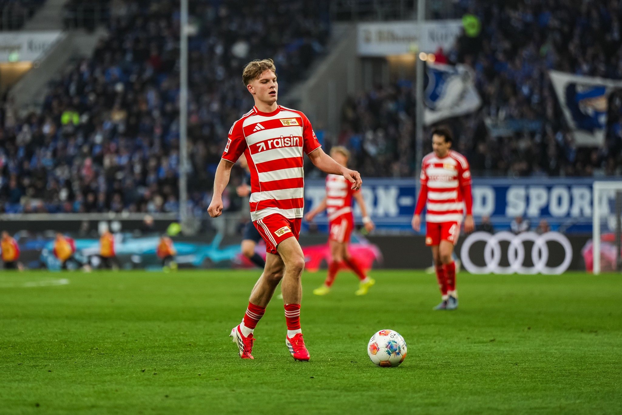 Spielszene mit Fußballspielern in rot-gestreiften Trikots auf dem Feld, im Hintergrund Fans und Vereinsbanner.