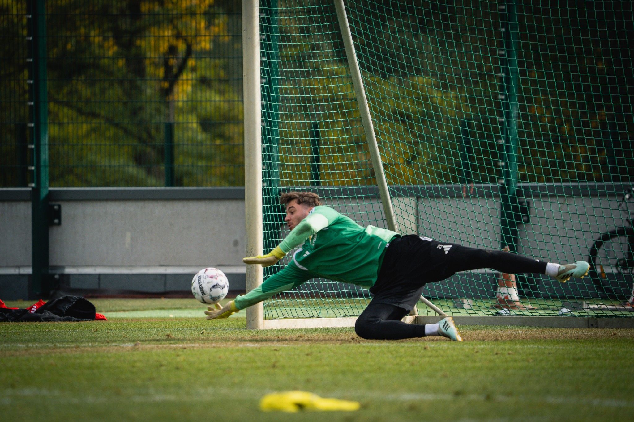 Ein Torwart in grüner Kleidung springt, um einen Fußball zu parieren, während er auf einem Rasenfeld trainiert.