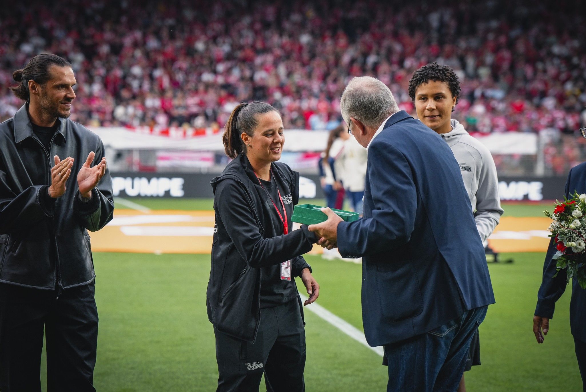 Eine Frau in Sportkleidung überreicht einem Mann bei einer Sportveranstaltung ein Geschenk, während Zuschauer im Hintergrund applaudieren.