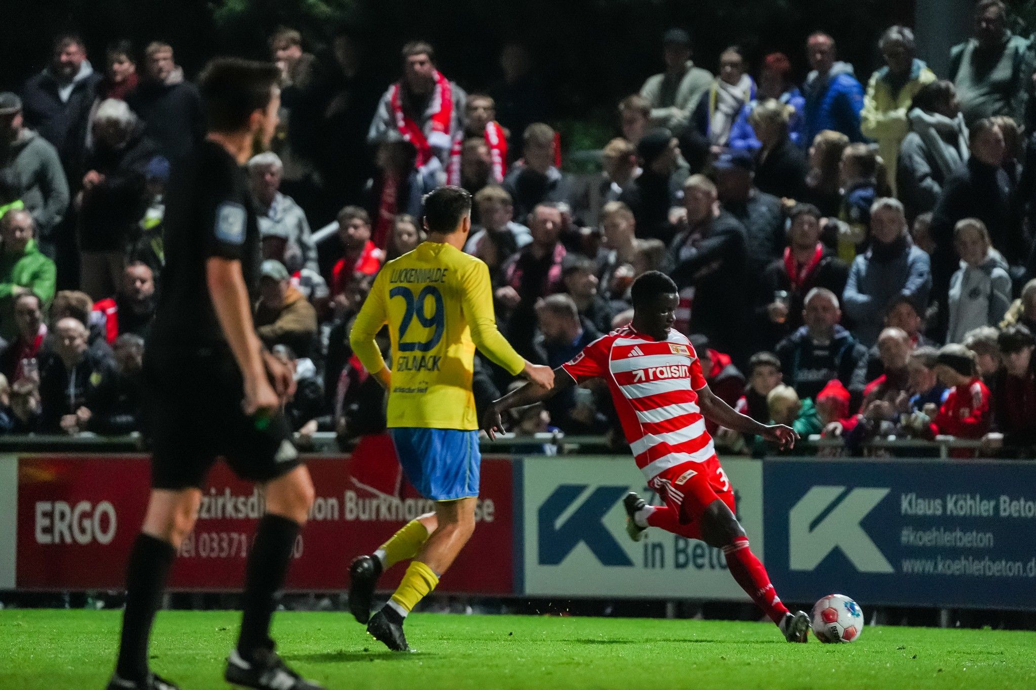 Ein Fußballspieler in rotem und weißem Trikot läuft mit dem Ball, während ein gegnerischer Spieler in gelbem Trikot hinter ihm ist. Zuschauer im Hintergrund.