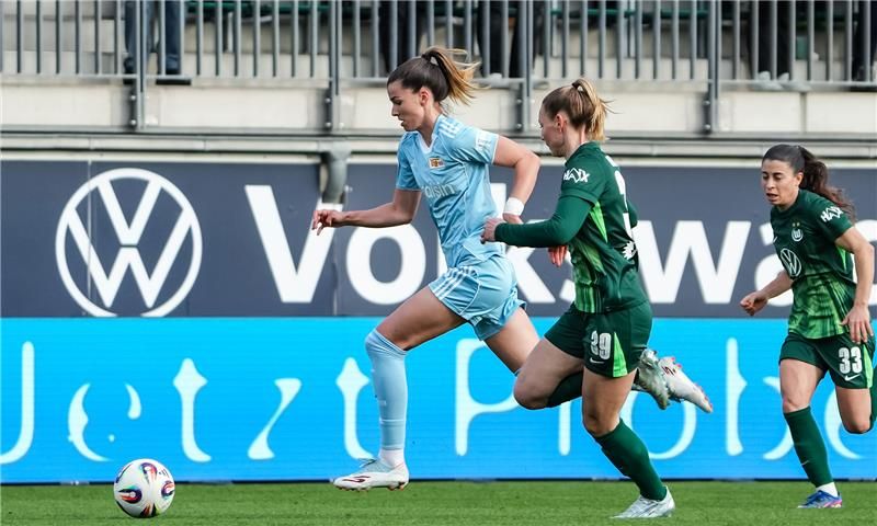 Spielszene: Eine Spielerin in blauen Trikots sprintet mit Ball, verfolgt von zwei Spielerinnen in grünen Trikots.