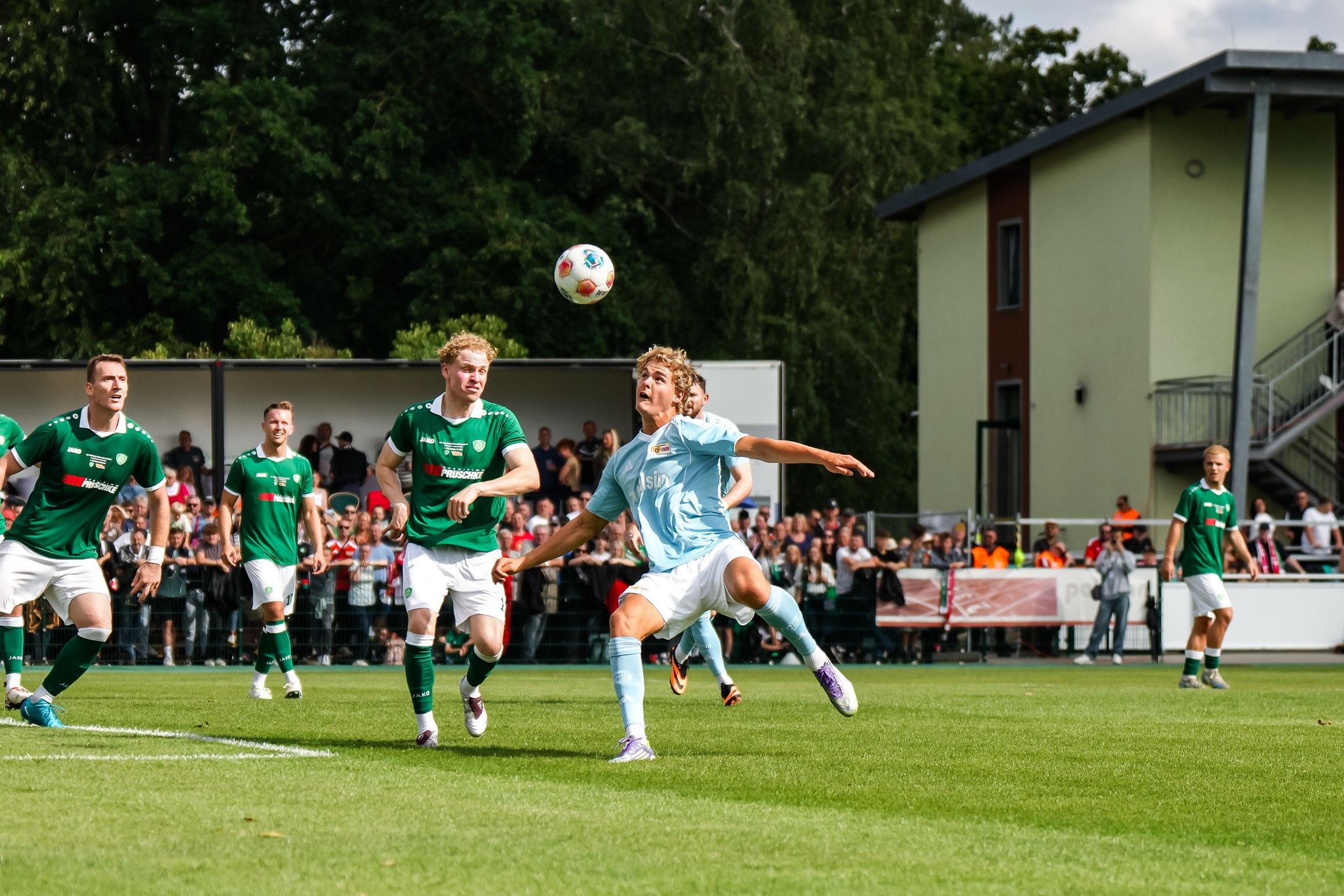 Ein Fußballspieler springt, um einen Ball während eines Spiels zu erreichen, umgeben von anderen Spielern und Zuschauern.