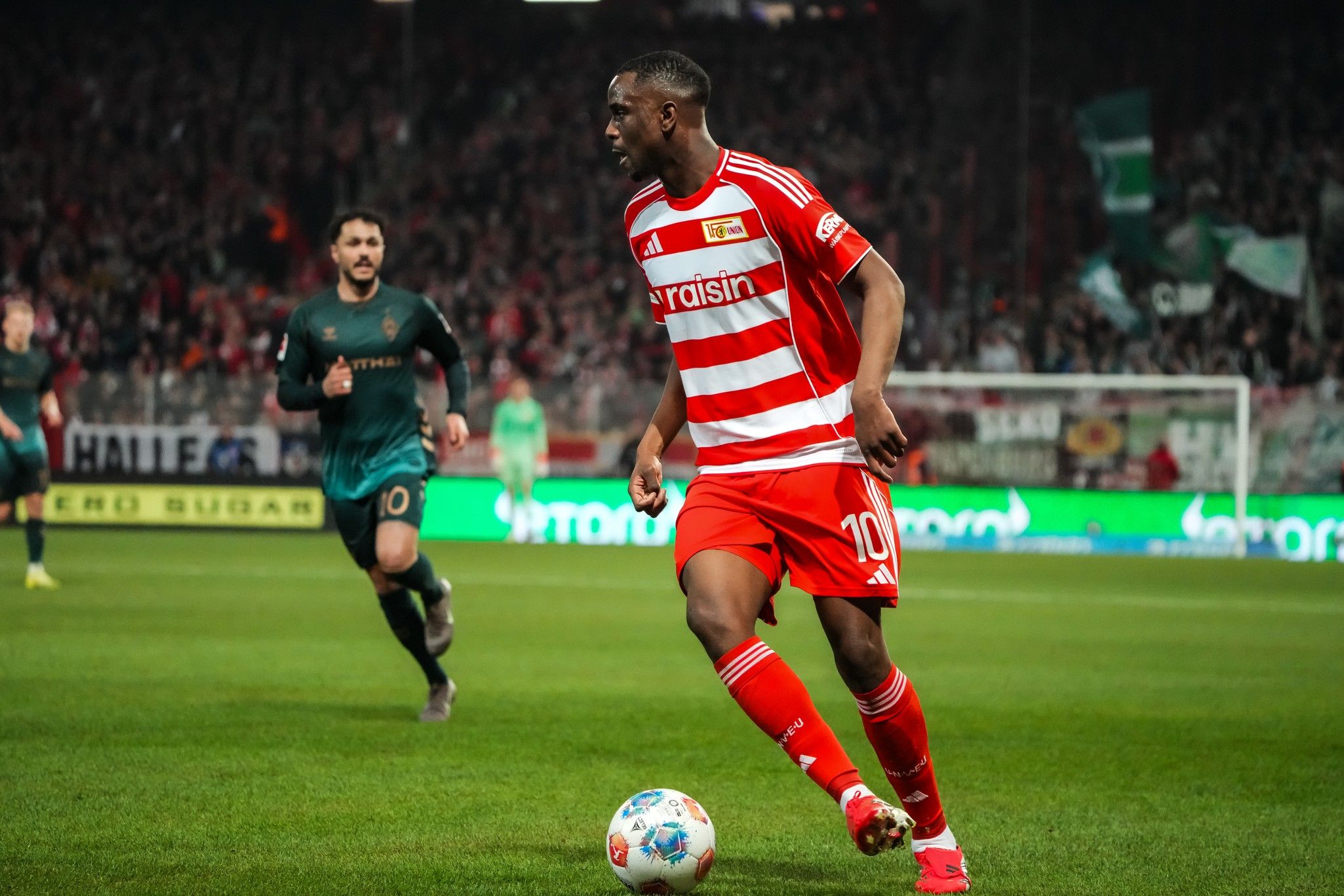 Ein Fußballspieler in rotem Trikot mit Streifen dribbelt den Ball auf einem Rasenplatz, während ein Gegner ihn verfolgt.