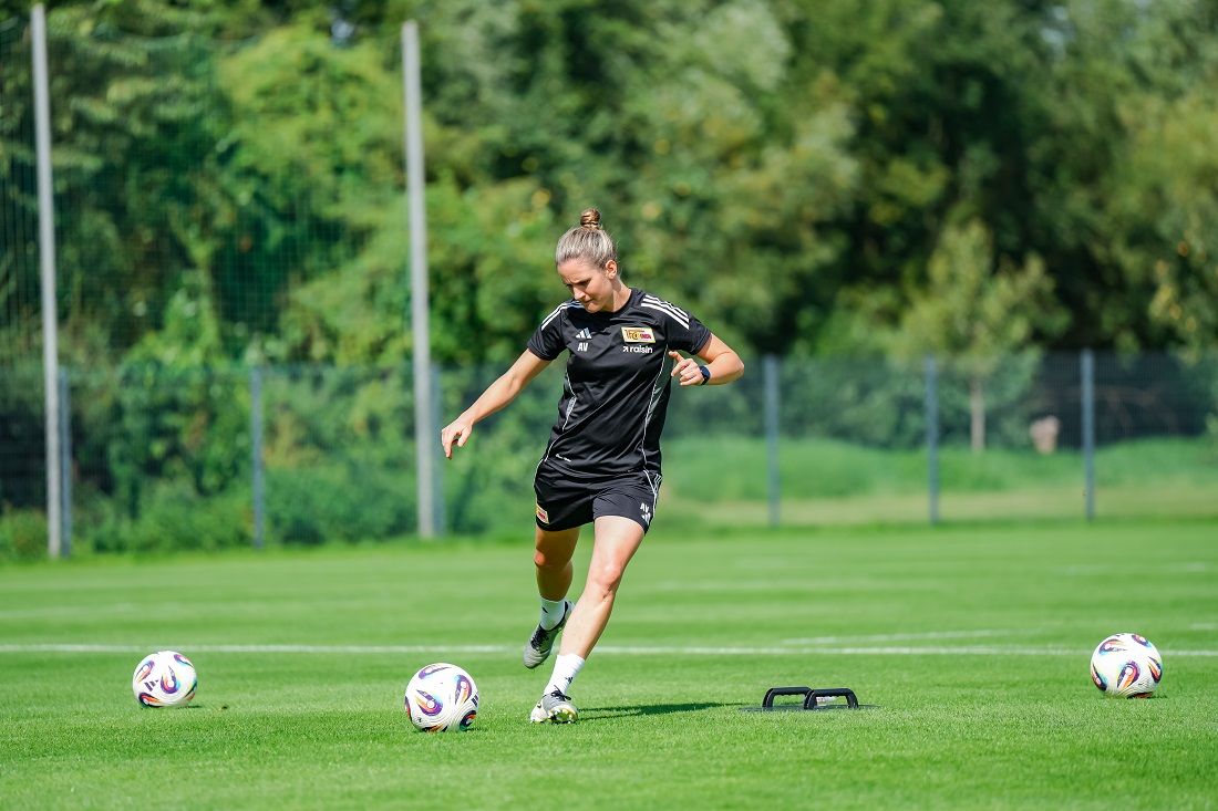 Eine Fußballspielerin in schwarzer Trainingskleidung trainiert auf einem grünen Platz mit mehreren Bällen.