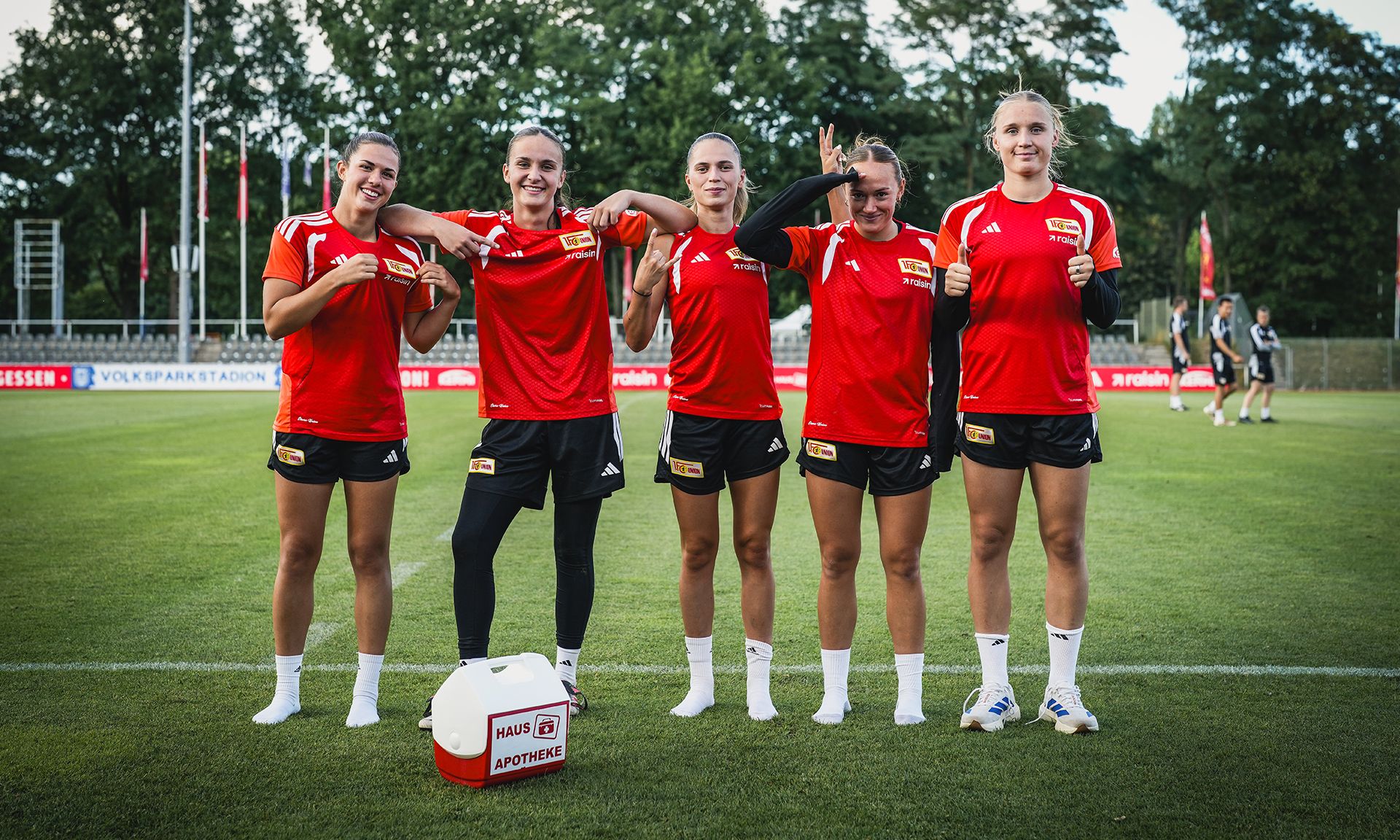 Fünf Sportlerinnen in roten Trikots stehen auf einem Fußballfeld, lächeln und posieren mit verschränkten Armen.