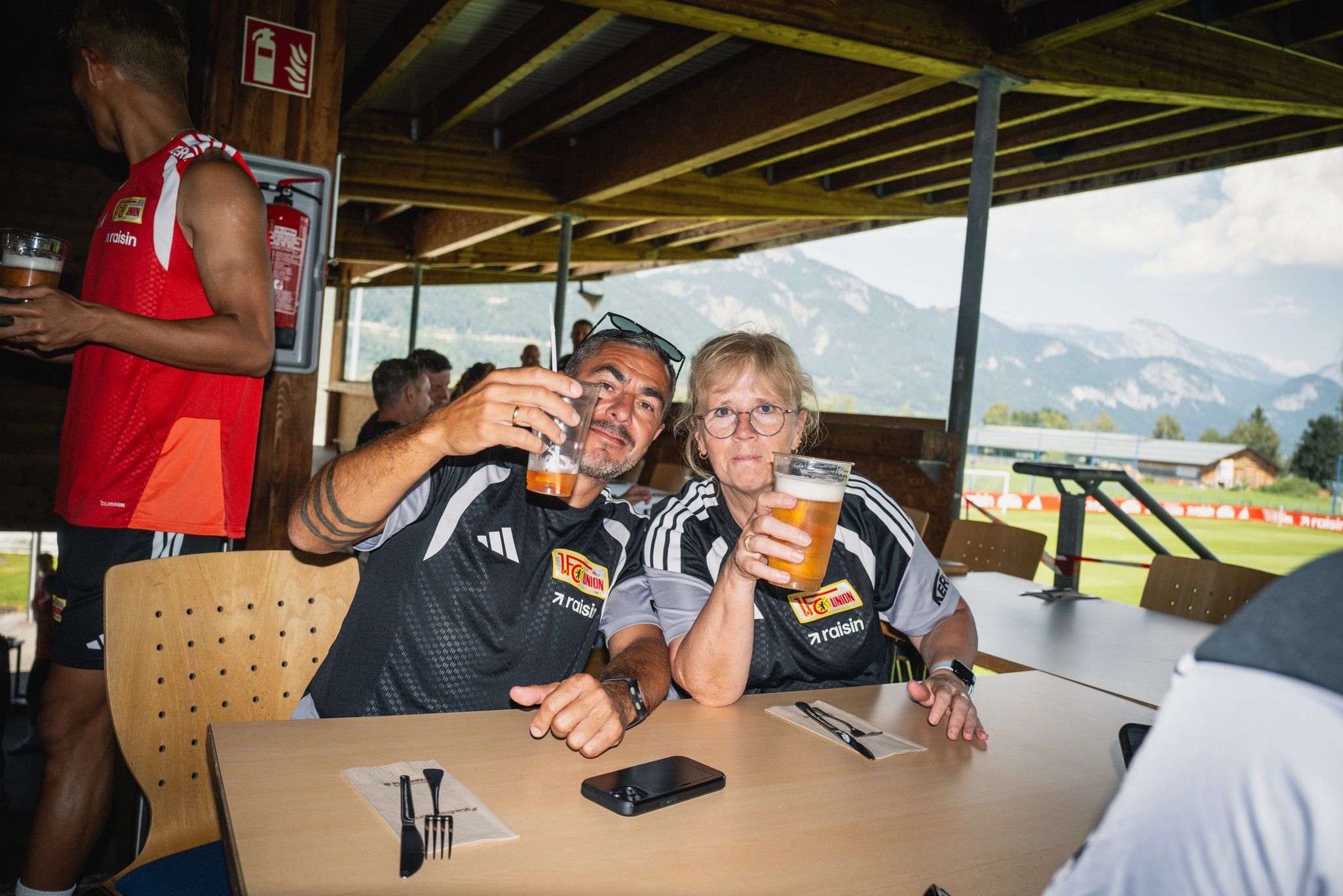 Zwei Personen sitzen an einem Tisch in einem Restaurant, halten Biergläser hoch und posieren für die Kamera.