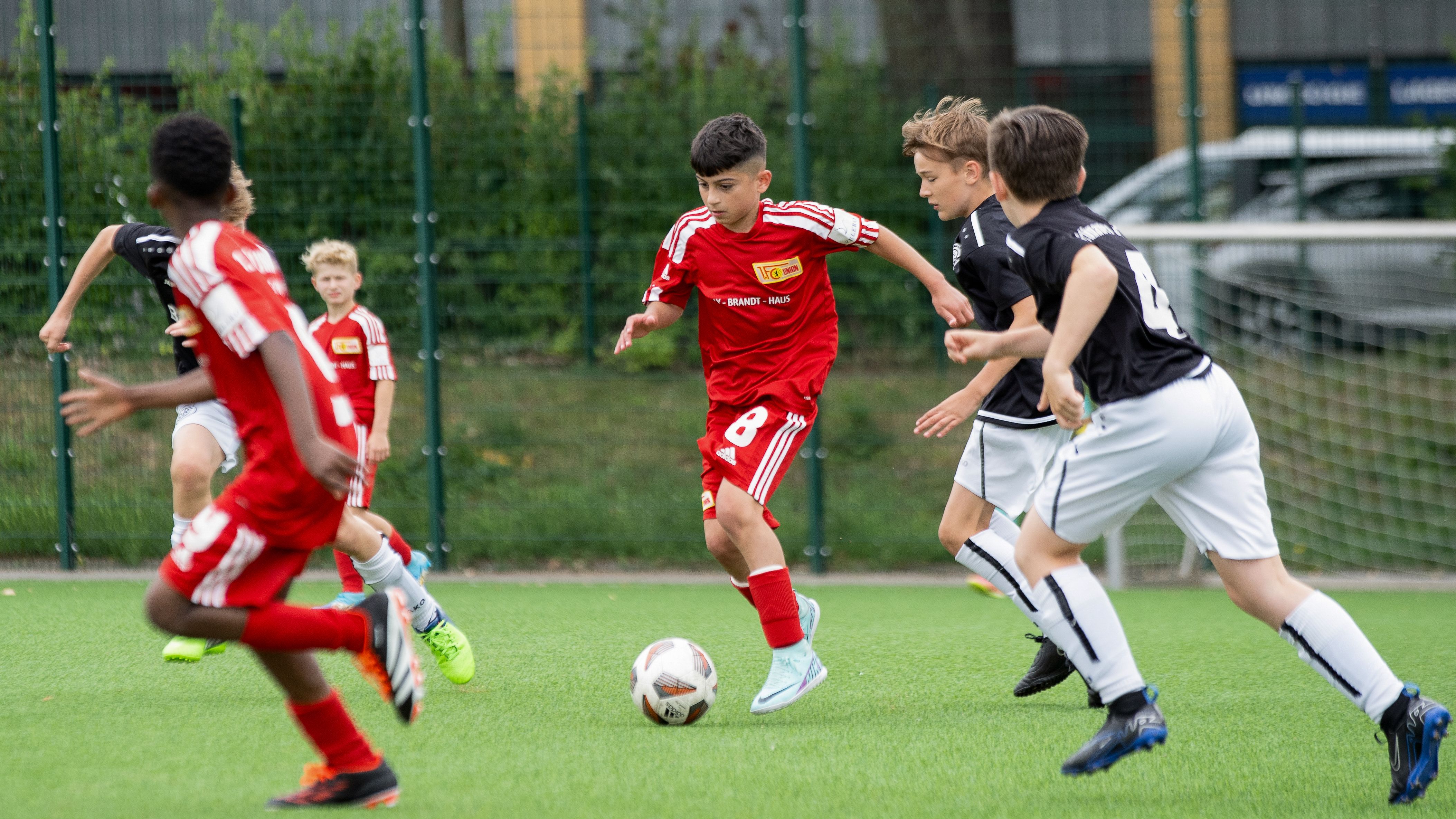 Ein Fußballspiel von Kindern, die in roten und schwarzen Trikots auf einem grünen Platz gegeneinander antreten.