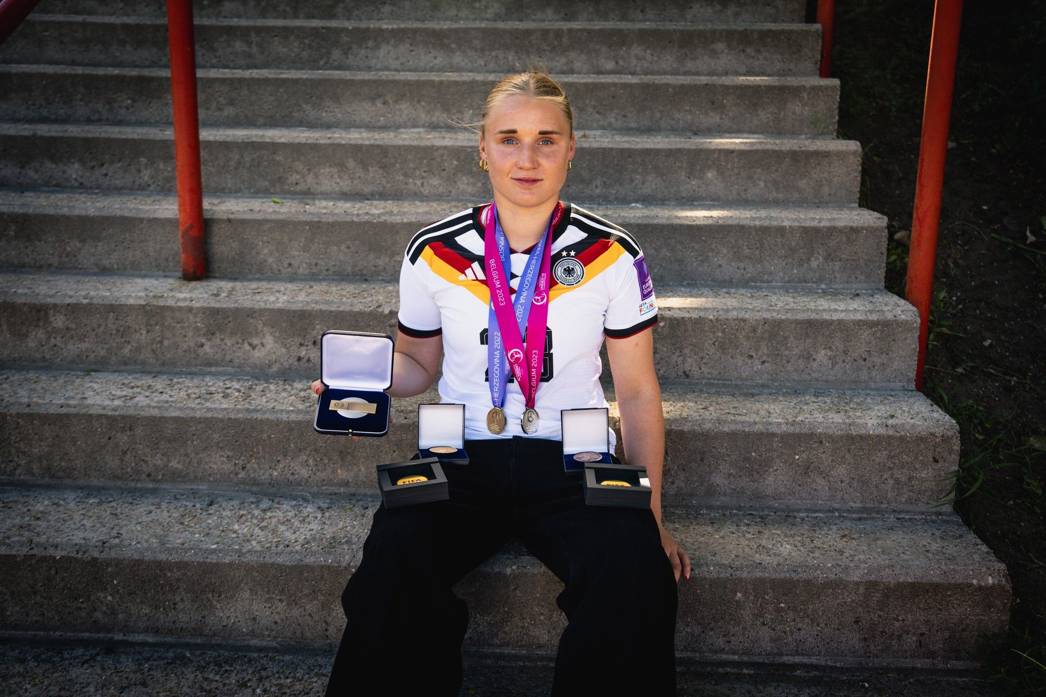 Eine Sportlerin in einem Trikot hält Medaillen und Auszeichnungen in der Hand, sitzt auf Treppenstufen.
