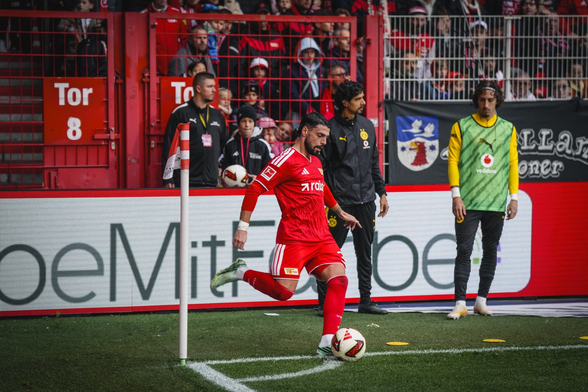 Spieler in rotem Trikot führt einen Eckstoß aus, während Trainer und Teamkollegen auf der Bank beobachten. Stadionatmosphäre im Hintergrund.