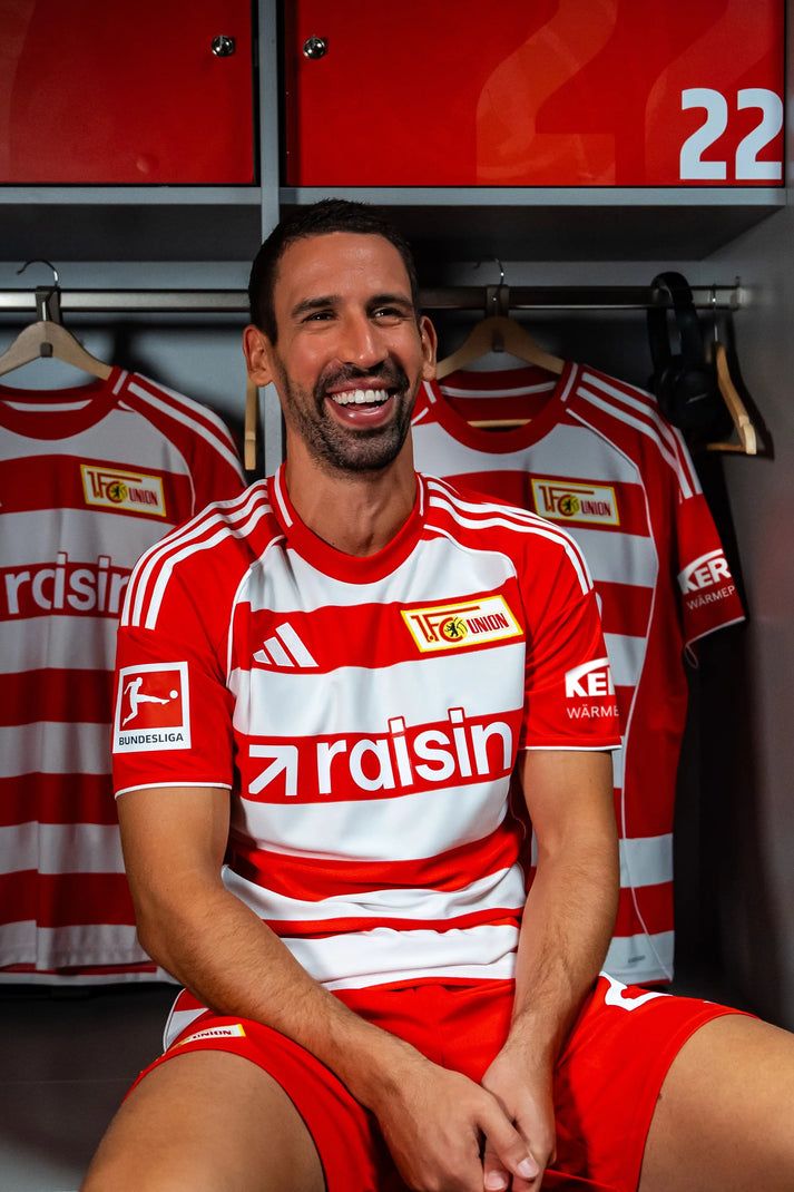Ein lachender Fußballspieler in einem roten Trikot mit weißen Streifen sitzt in einer Umkleidekabine. Hinter ihm hängen weitere Trikots.