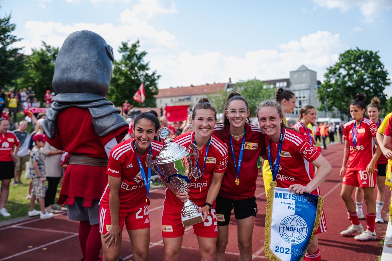 Vier Spielerinnen in roten Fußballtrikots posieren mit einem Pokal und einer Urkunde. Im Hintergrund sind jubelnde Fans sichtbar.