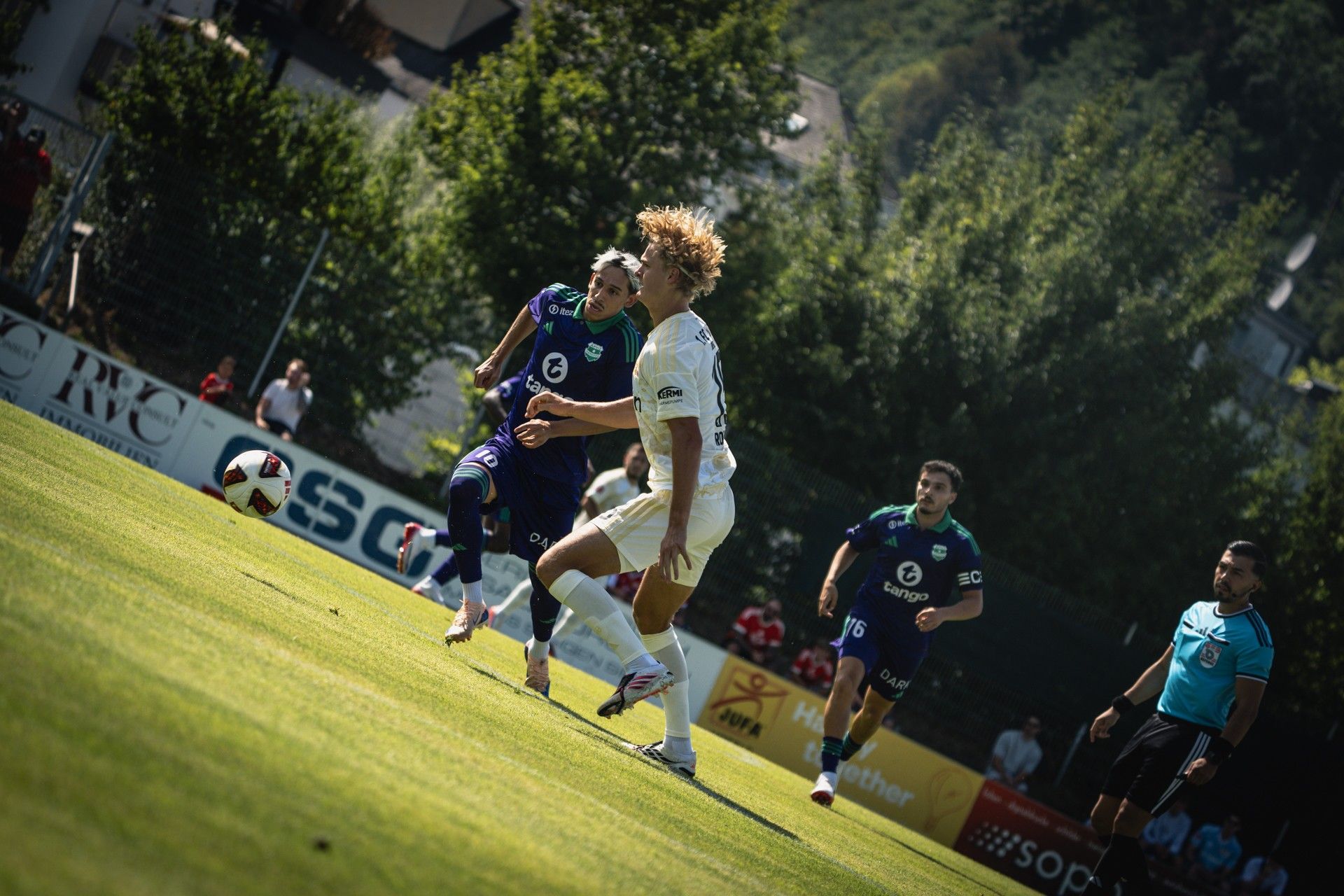 Zwei Fußballspieler laufen auf einem Spielfeld, während ein dritter Spieler im Hintergrund zuschaut. Sonne scheint, Bäume im Hintergrund.