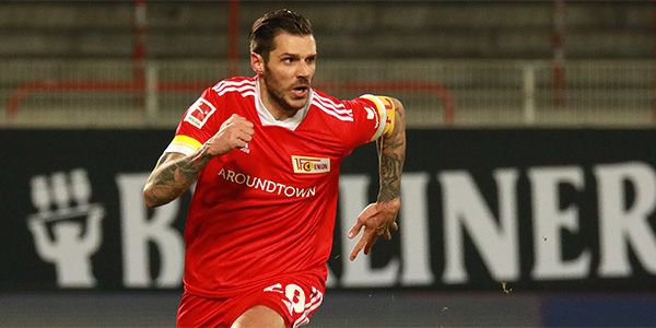 A soccer player in a red jersey sprints across the field with a determined expression.