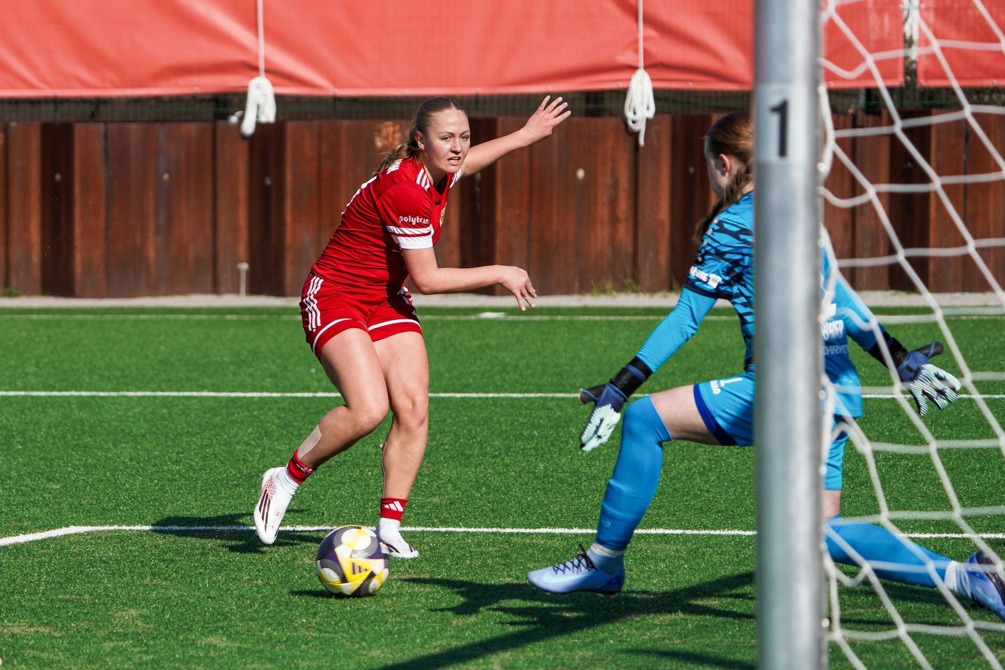 Eine Fußballspielerin in rot versucht, die gegnerische Torfrau in blau zu überwinden, während sie auf das Tor zuläuft.