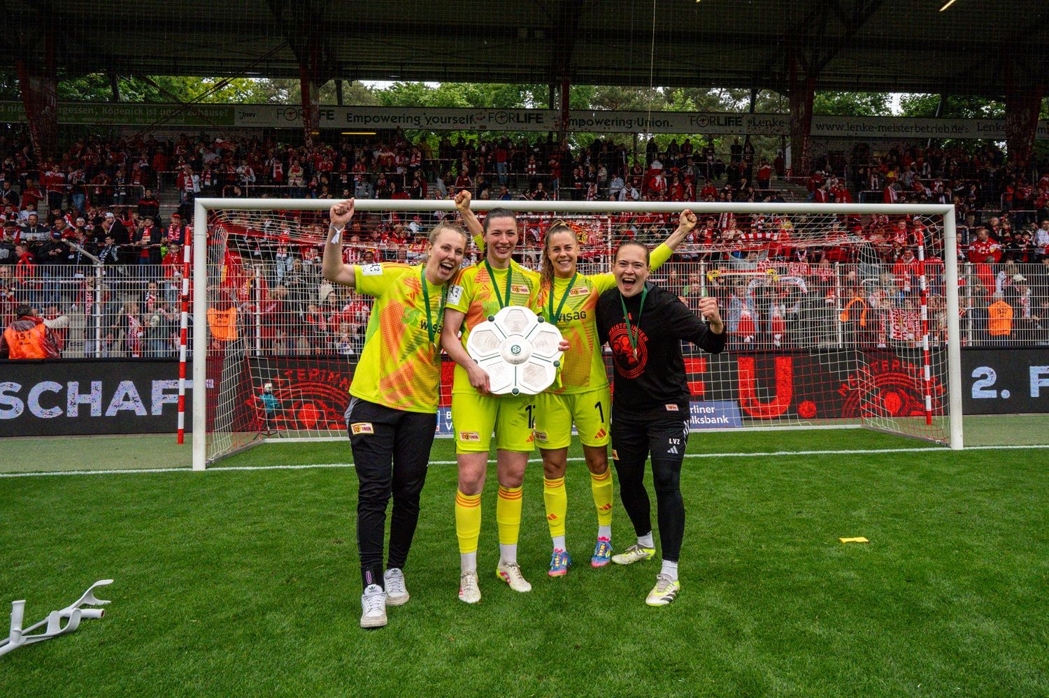Vier Fußballspielerinnen feiern auf dem Platz, halten eine Trophäe hoch und lächeln vor jubelndem Publikum.