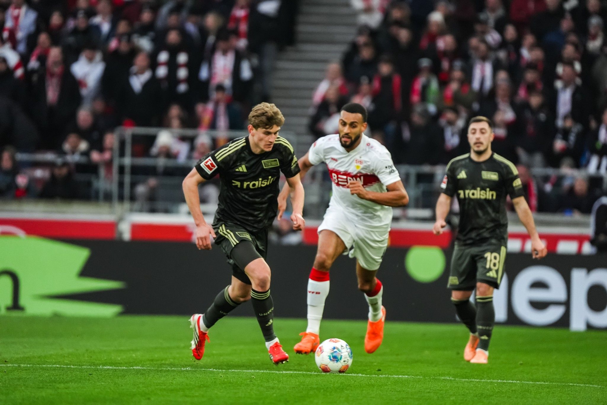 Zwei Fußballspieler in Aktion auf dem Spielfeld, ein Spieler in schwarzem Trikot dribbelt, während ein Spieler in weißem Trikot verfolgt.