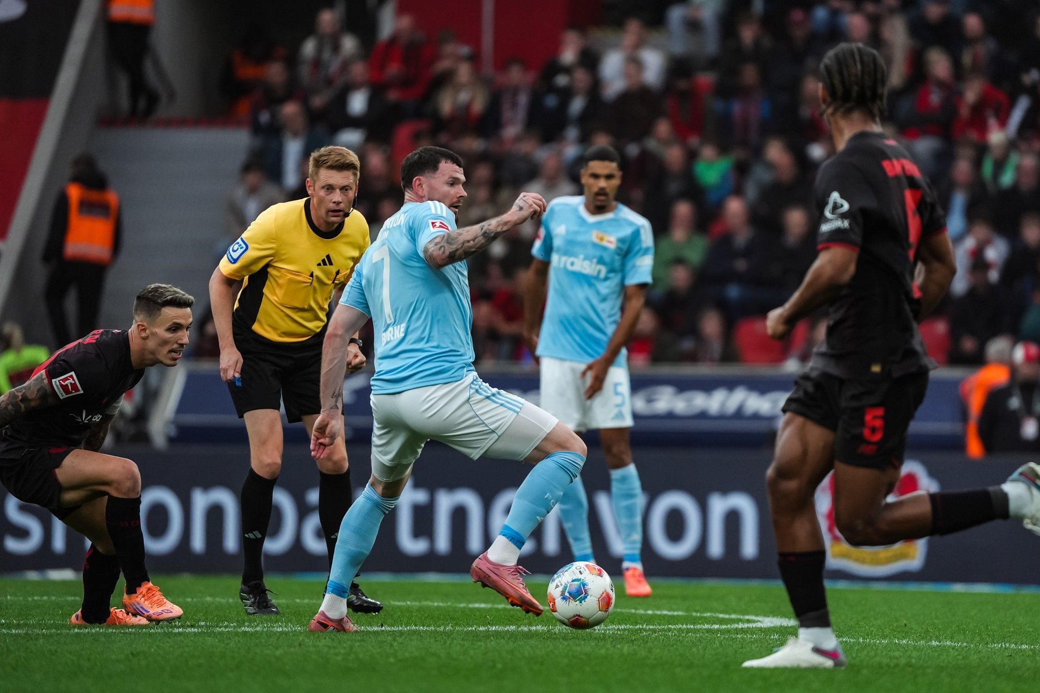 Ein Fußballspiel im Gange, mit Spielern in blauem und schwarzem Trikot, einem Schiedsrichter und angespannter Atmosphäre.