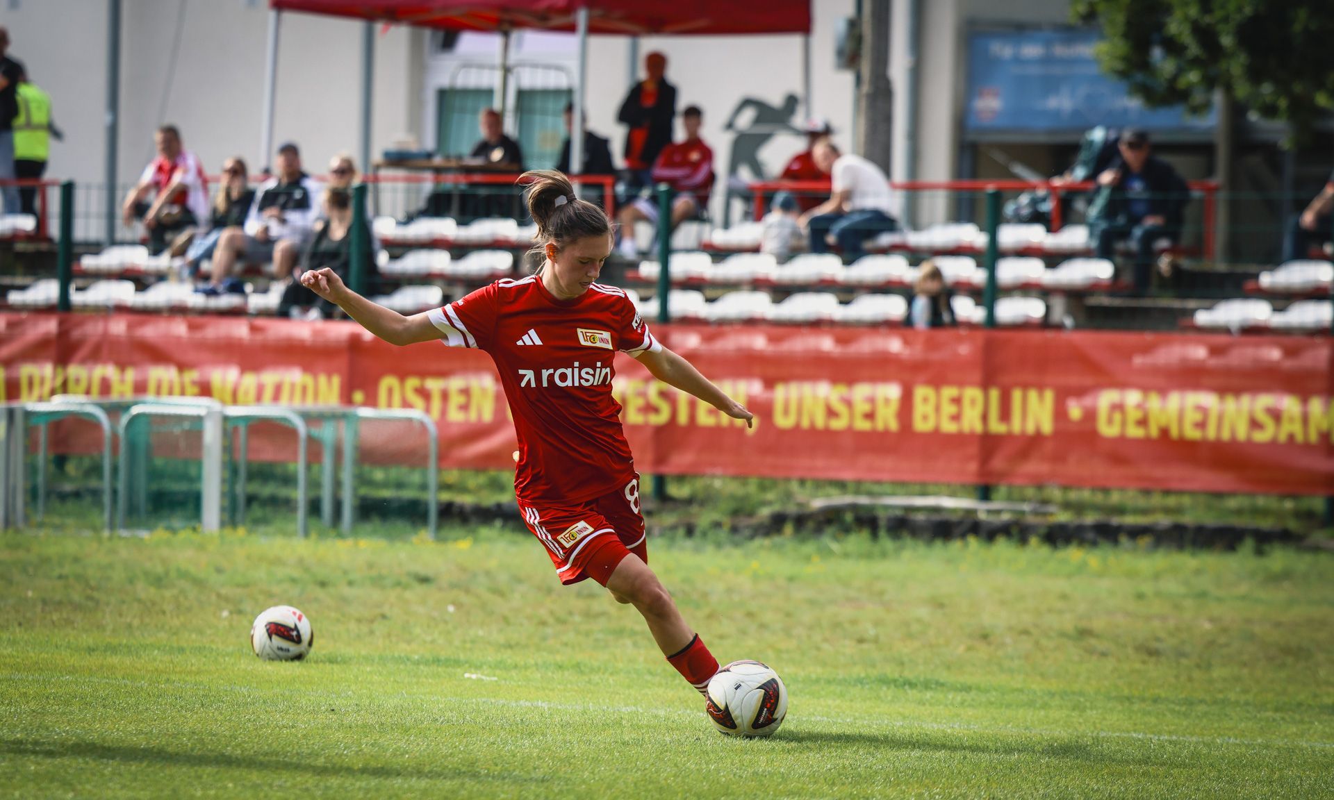 Eine Fußballspielerin in roter Ausrüstung tritt den Ball auf einem Spielfeld mit Zuschauern im Hintergrund.