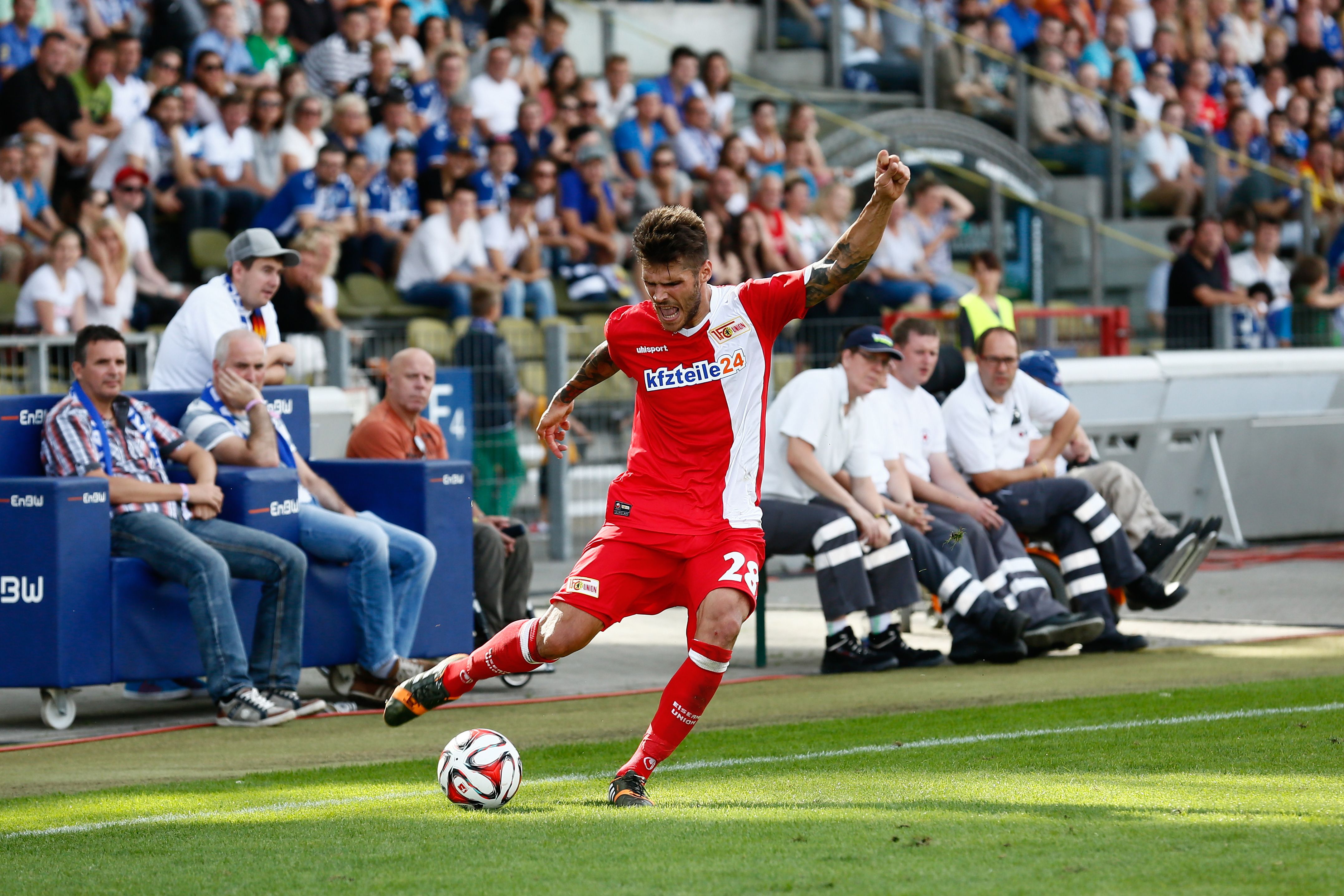 Ein Fußballspieler in rotem Trikot tritt den Ball während eines Spiels an, im Hintergrund jubelnde Fans und Zuschauer.
