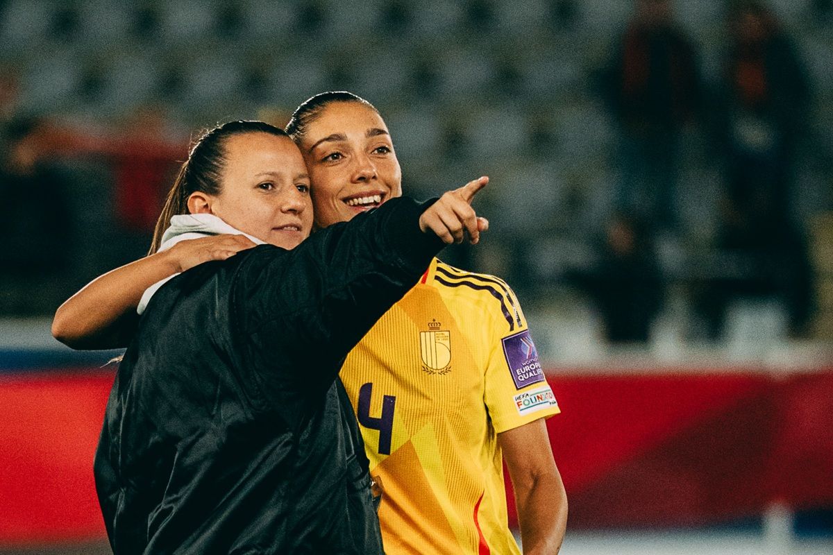 Zwei Frauen umarmen sich auf dem Fußballfeld, eine zeigt mit dem Finger in eine Richtung und lächelt.