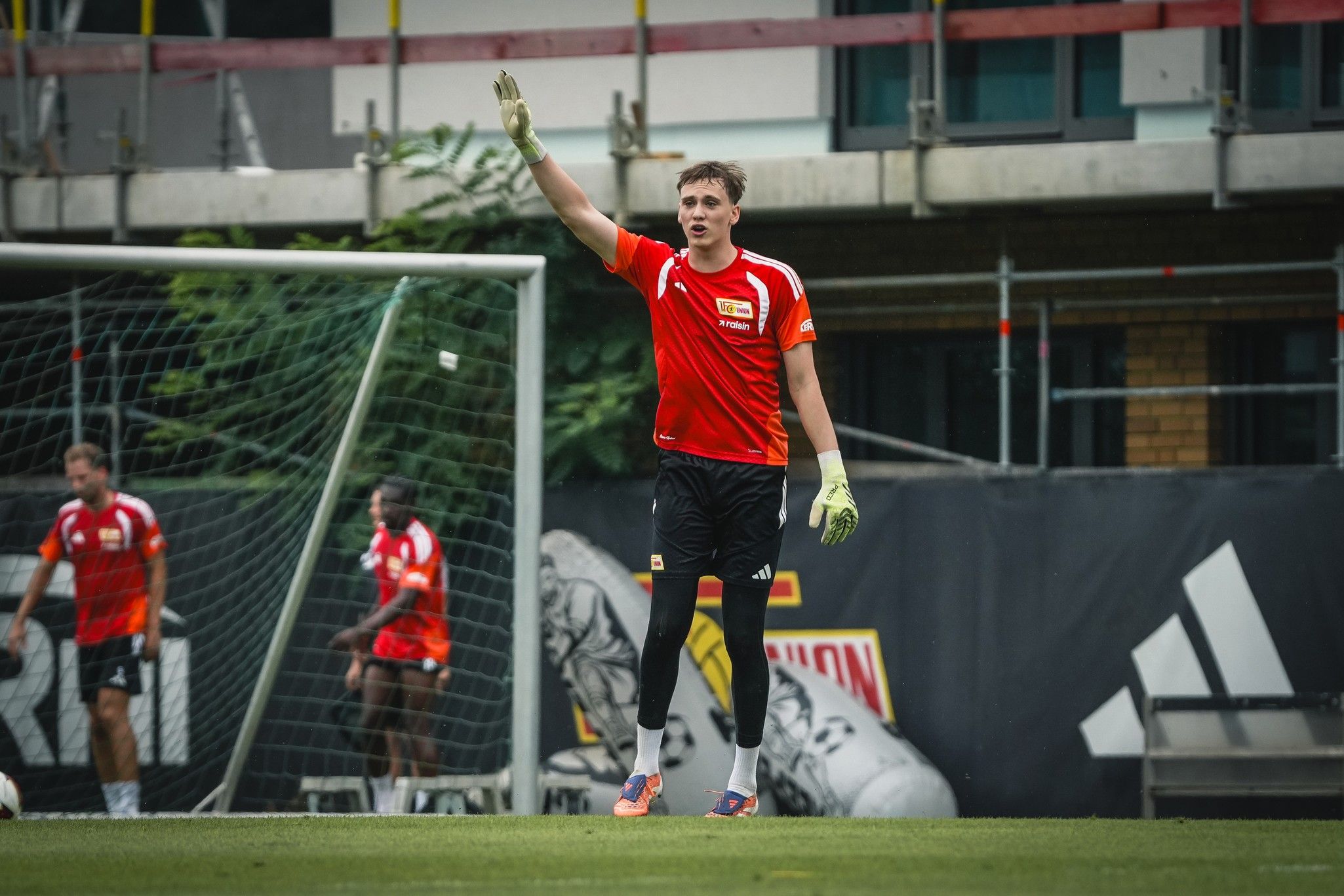 Torwart mit Handschuhen hebt die Hand während des Trainings auf dem Fußballplatz, im Hintergrund Spieler in roten Trikots.