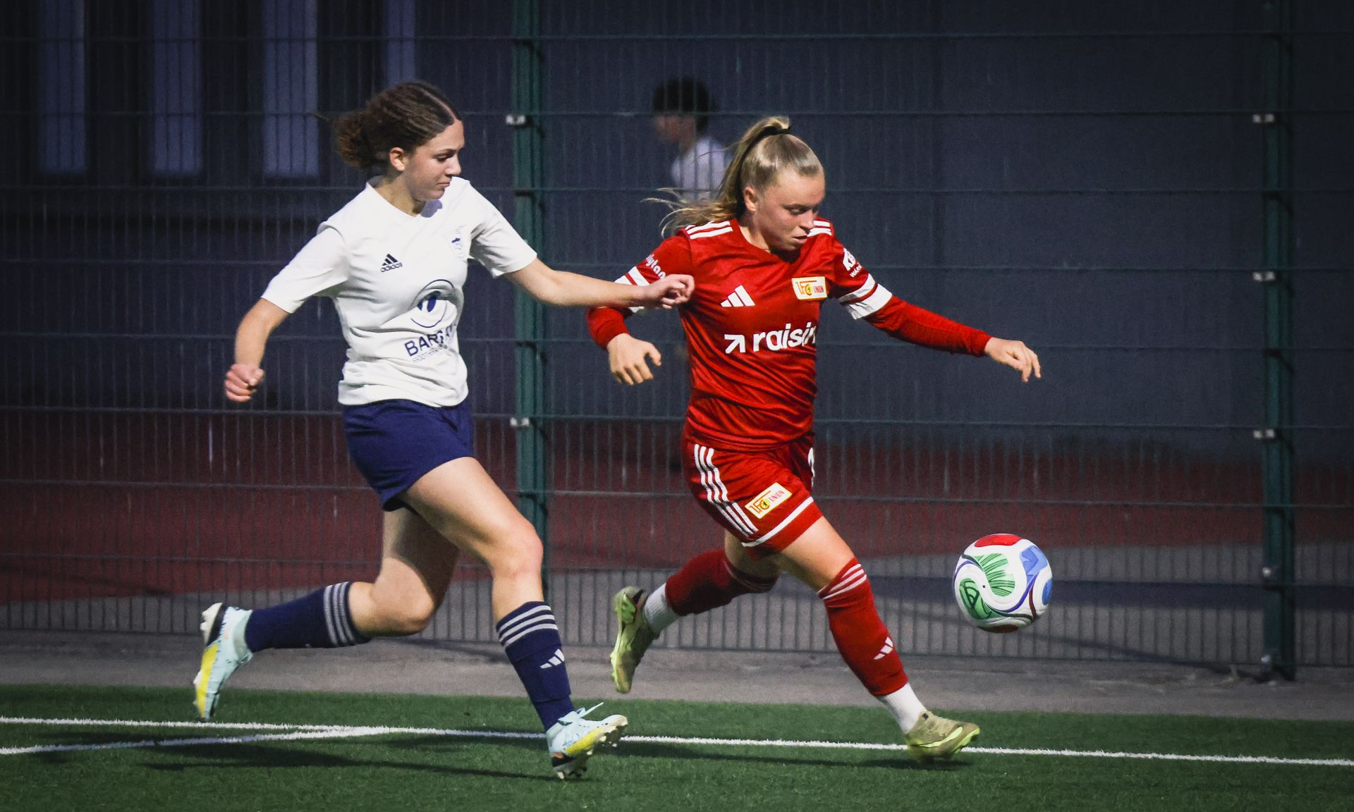Zwei Frauen im Fußballspiel, eine in rotem Trikot spielt den Ball, die andere in weißem Trikot versucht, ihn zu stoppen.