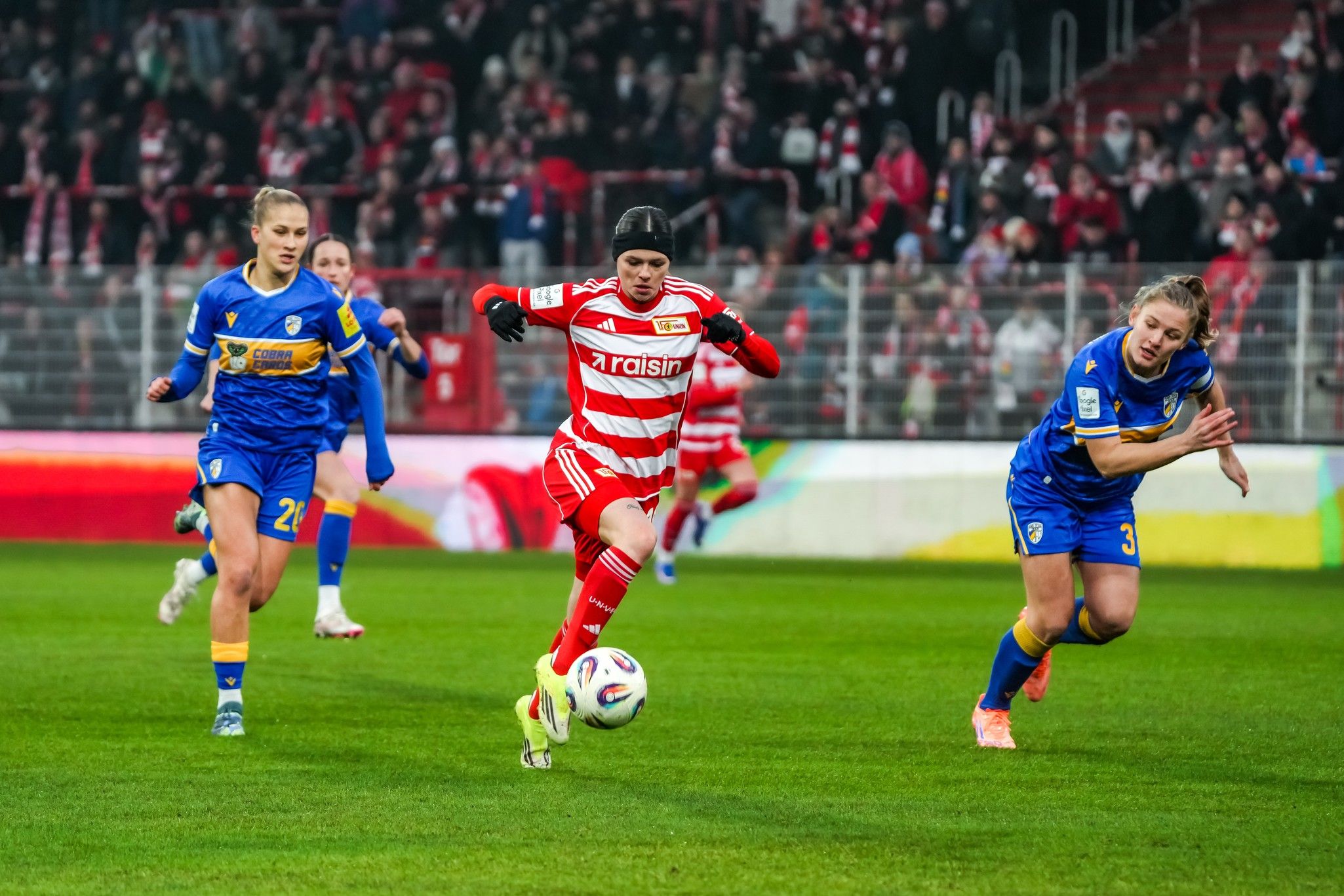 Eine Spielerin in roten und weißen Streifen dribbelt den Ball, während zwei Spielerinnen in blauen Trikots verfolgen.