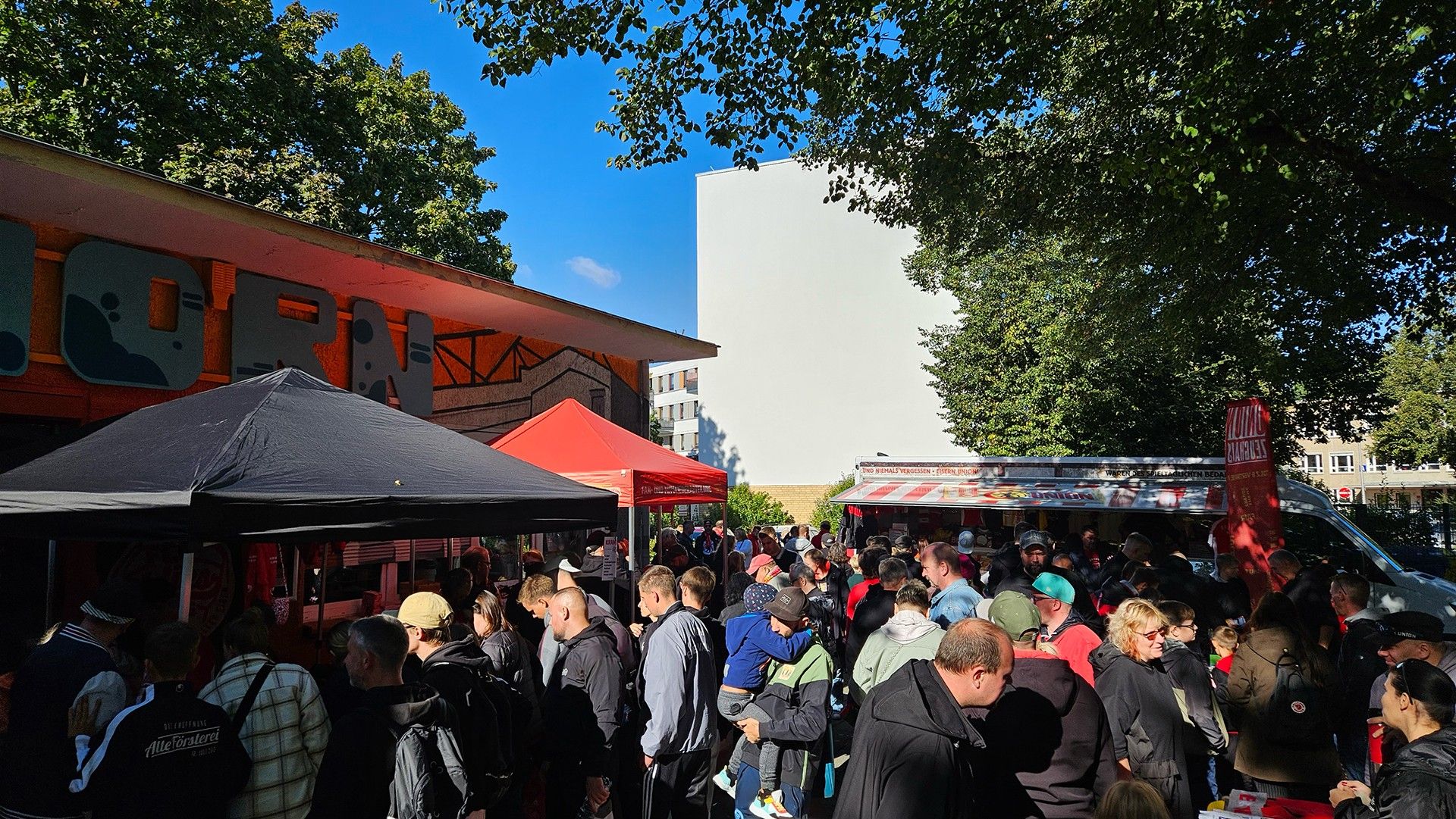 Menschenmenge vor einem Gebäude mit Veranstaltungsständen und Essenswagen bei sonnigem Wetter.
