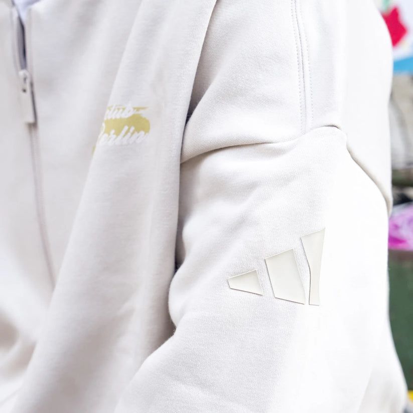 Nahaufnahme eines hellen Sweatshirts mit einem Adidas-Logo auf dem Ärmel und einem weiteren Logo auf der Vorderseite.