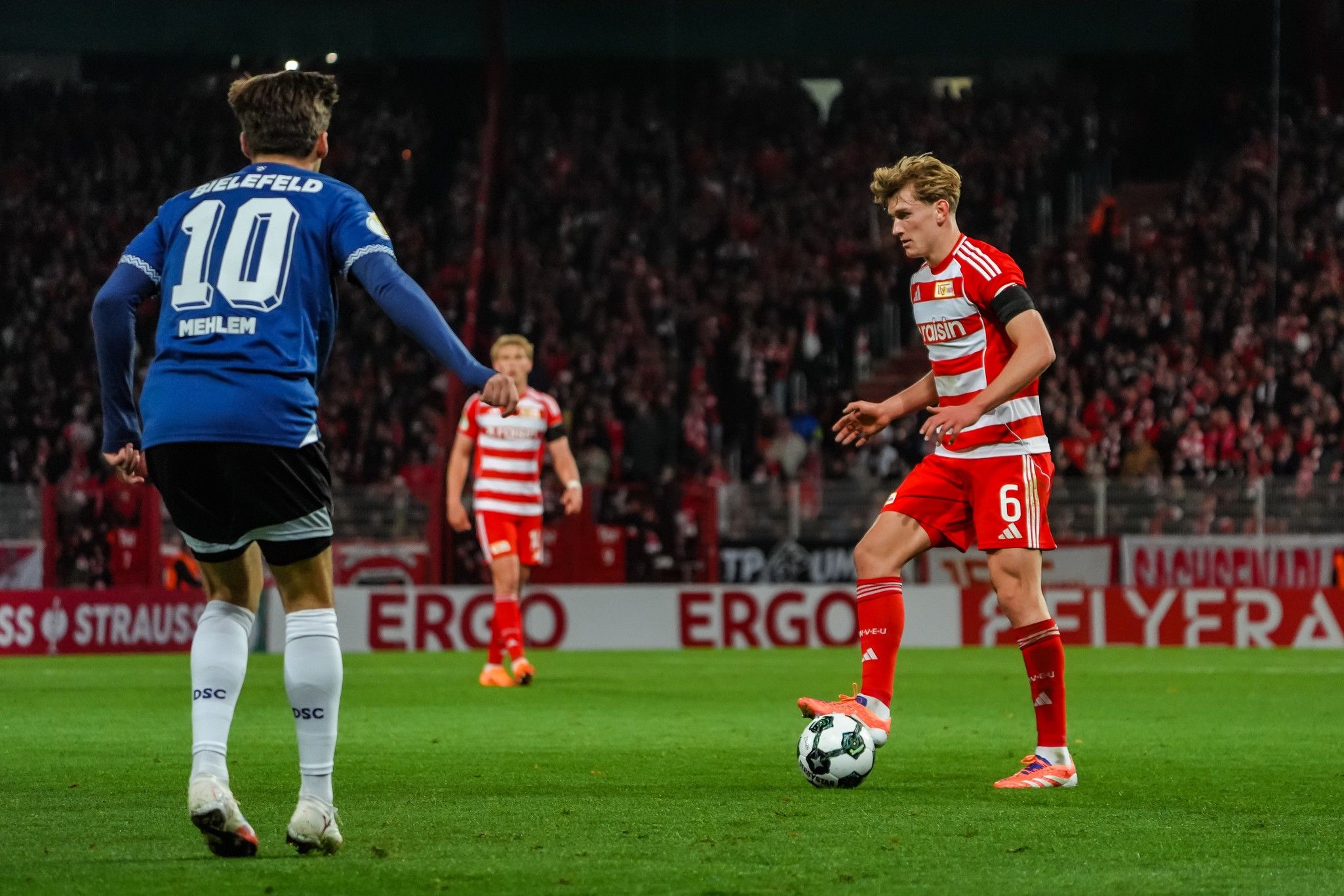 Spielszene eines Fußballspiels mit zwei Spielern, einem in blauer und einem in roter Uniform, vor einem belebten Stadionhintergrund.