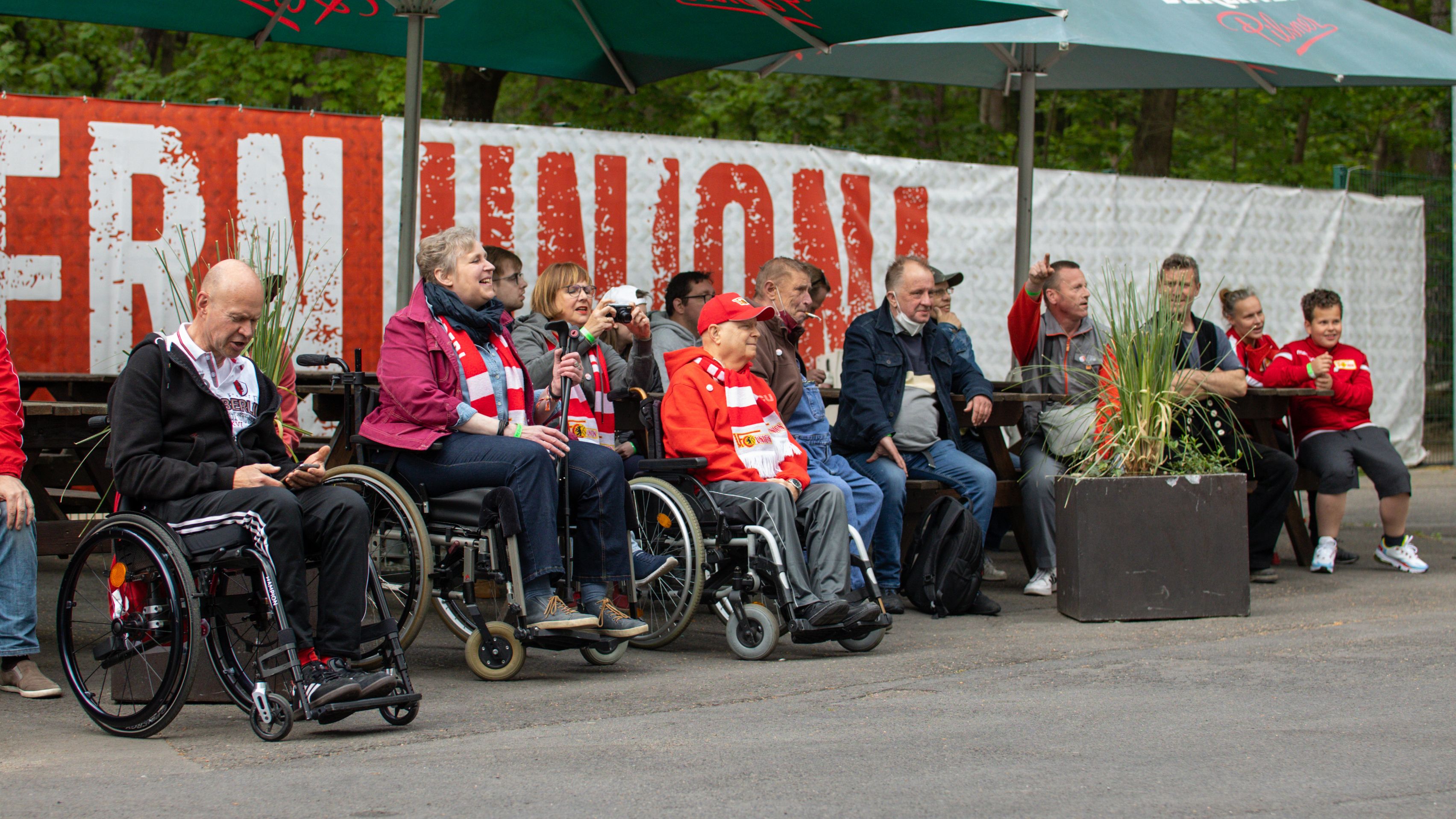 Grupppe von Menschen, einige im Rollstuhl, sitzen unter Markisen und verfolgen ein Event im Freien.