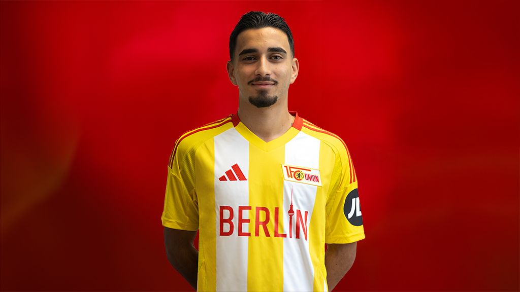 Ein junger Mann in einem gelben Fußballtrikot mit roten und weißen Akzenten steht vor einem roten Hintergrund.
