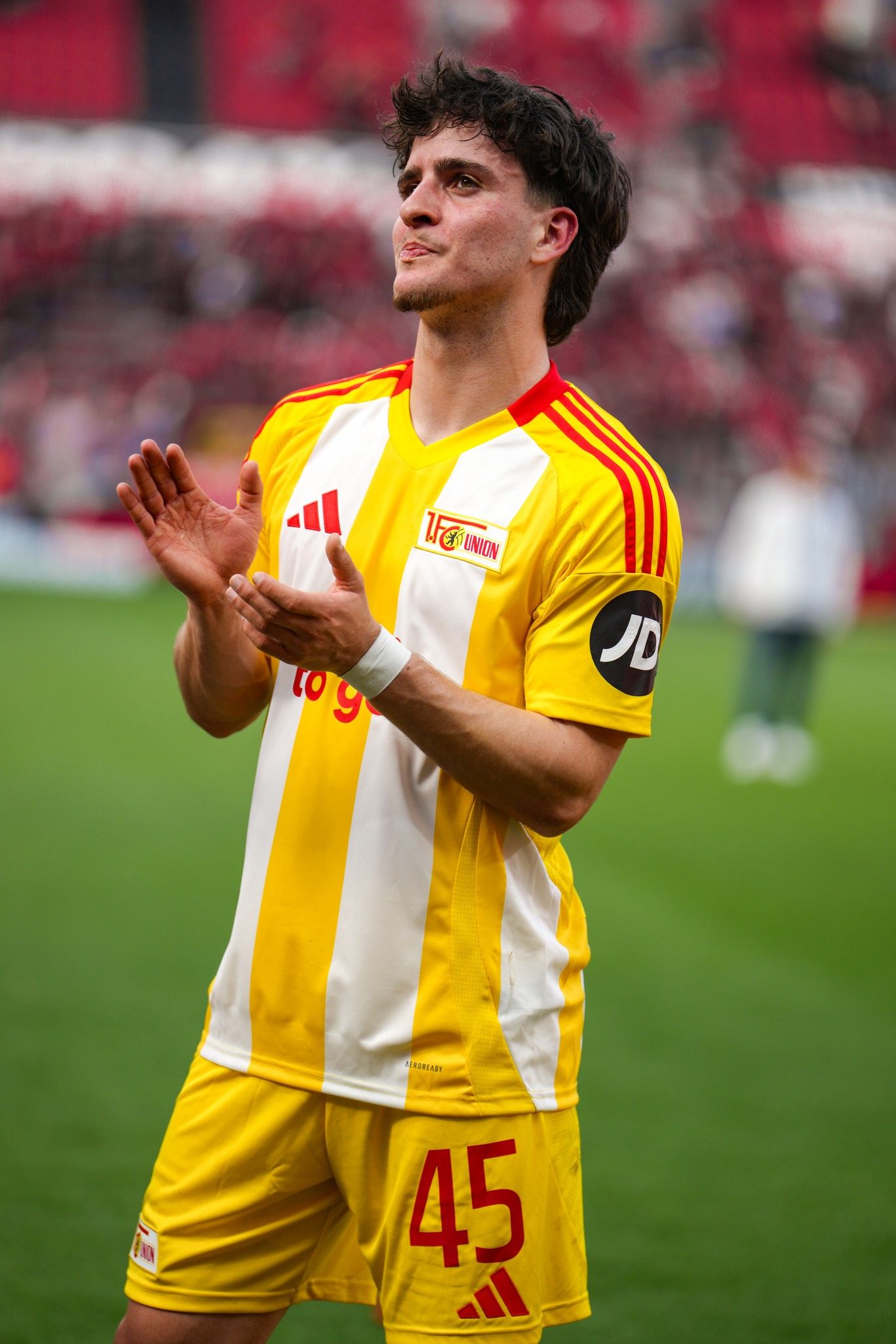 Ein Fußballspieler in gelb-rot gestreiftem Trikot applaudiert, mit der Nummer 45 auf dem Rücken, auf dem Spielfeld.