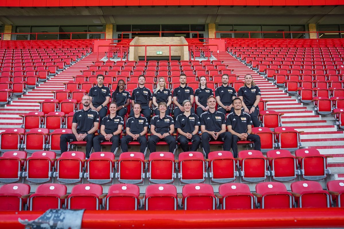 Eine Gruppe von 20 Menschen in schwarzen Outfits sitzt auf einer roten Tribüne in einem Stadion.