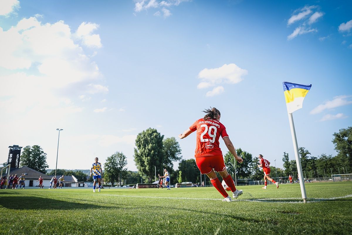 Eine Fußballspielerin in rotem Trikot spielt einen Eckball auf einem grünen Fußballfeld unter blauem Himmel.