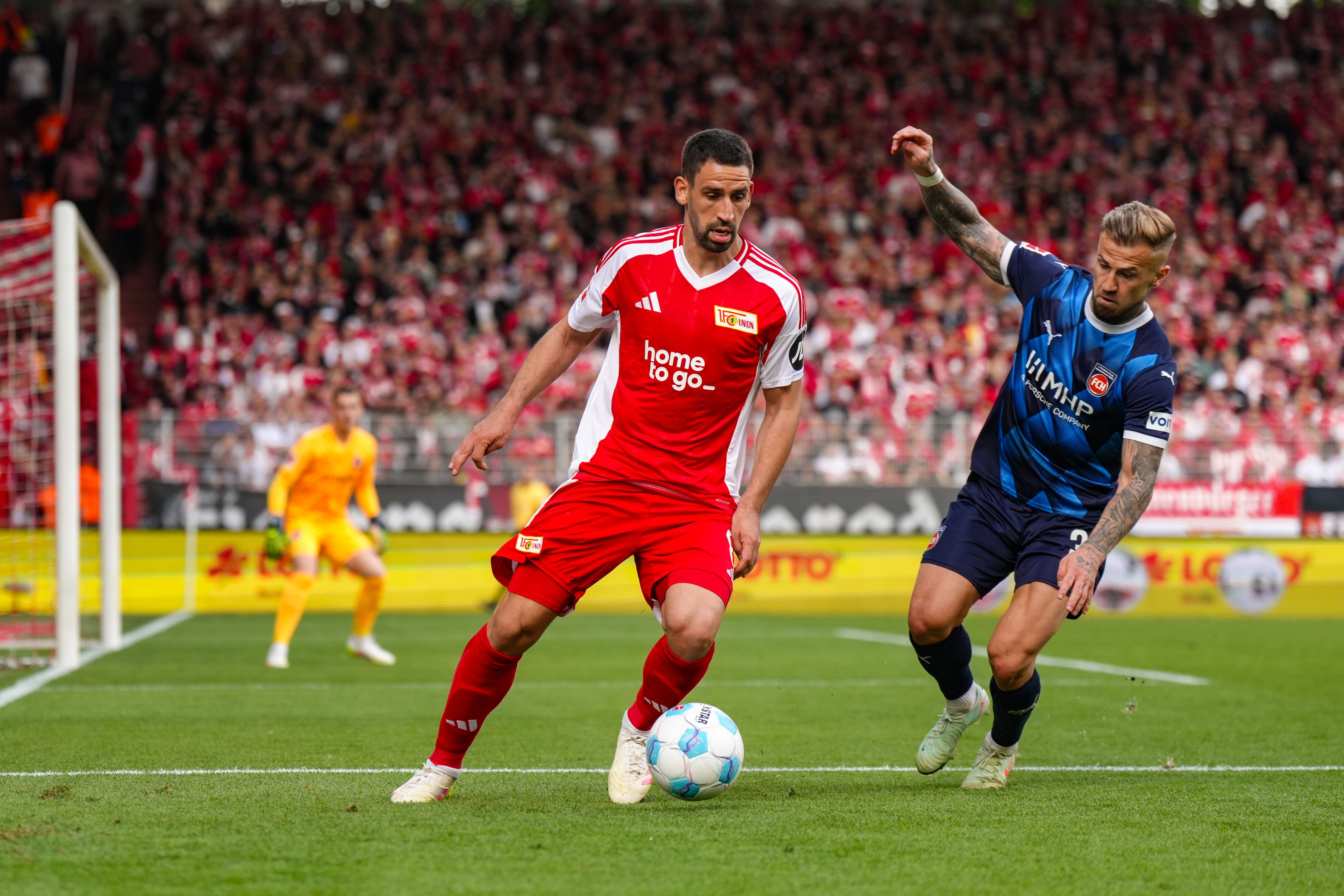Ein Fußballspieler in rotem Trikot dribbelt den Ball, während ein gegnerischer Spieler in blauem Trikot ihm folgt.