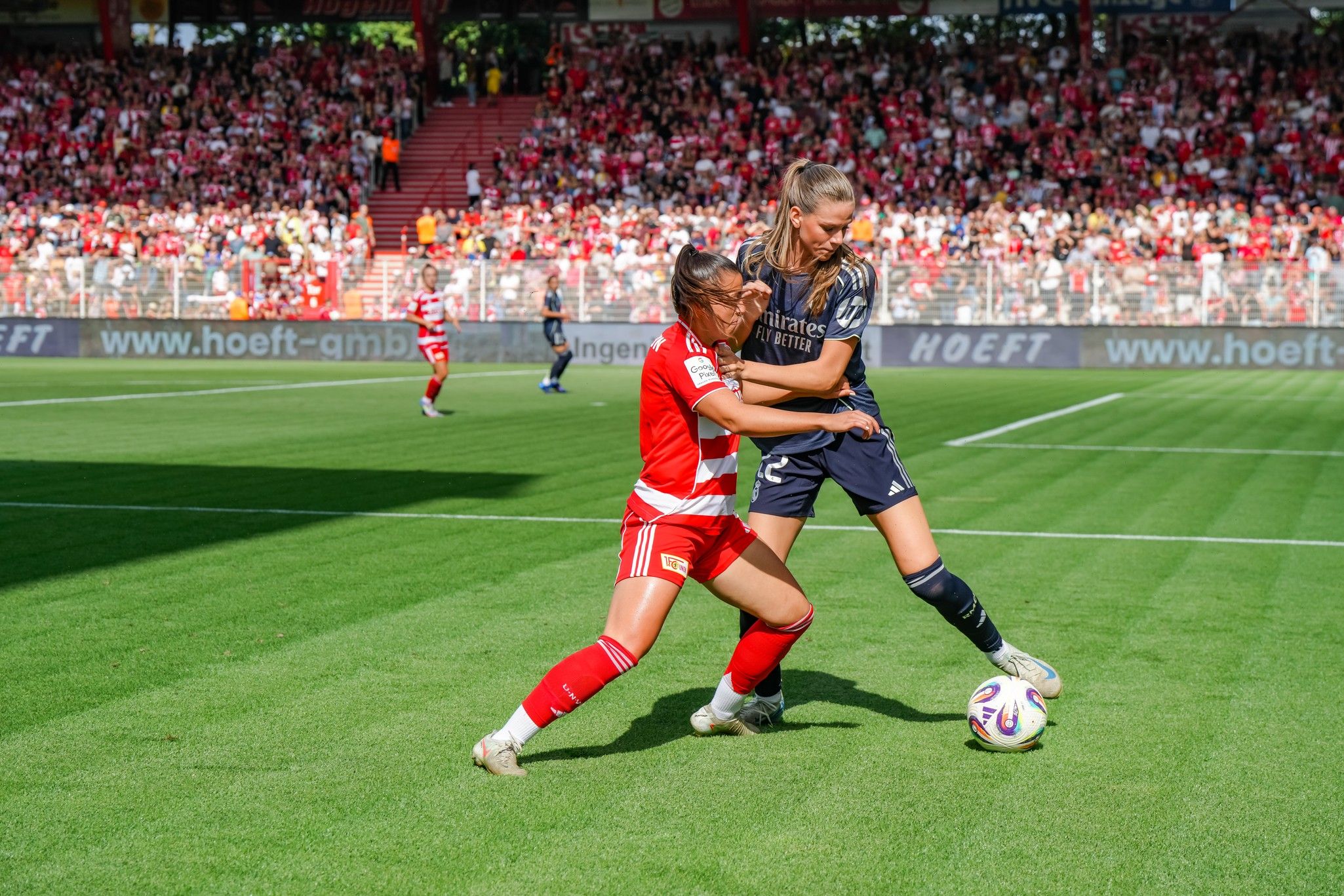 Zwei Fußballspielerinnen kämpfen um den Ball auf einem Rasenplatz während eines Spiels in einem Stadion.