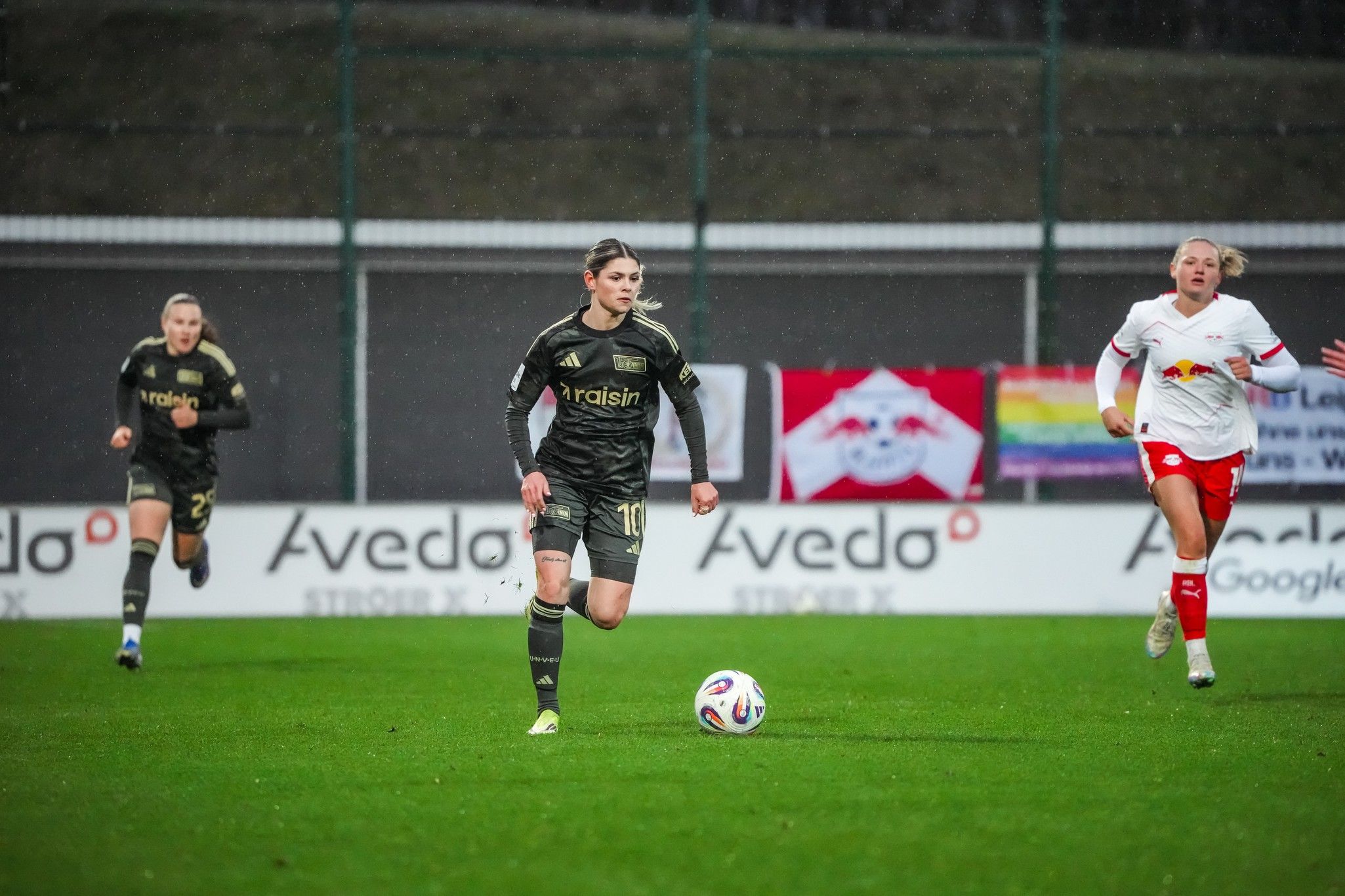 Eine Fußballspielerin dribbelt den Ball auf einem nassen Spielfeld, während zwei weitere Spielerinnen im Hintergrund folgen.