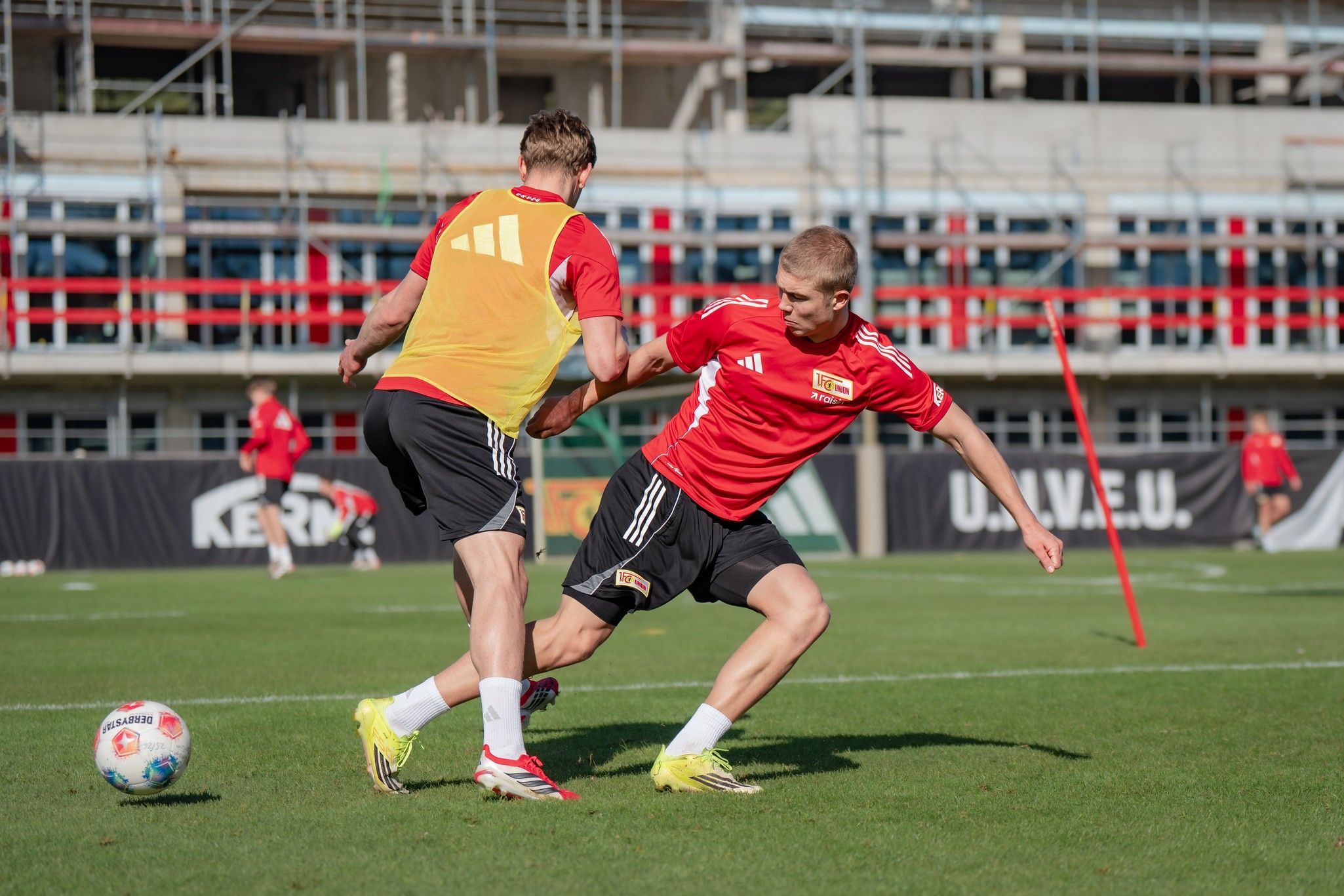 Zwei Fußballspieler in Training, einer mit gelbem Trainingsshirt, der andere in rotem Shirt, beim Dribbeln und Abwehrspiel auf dem Platz.