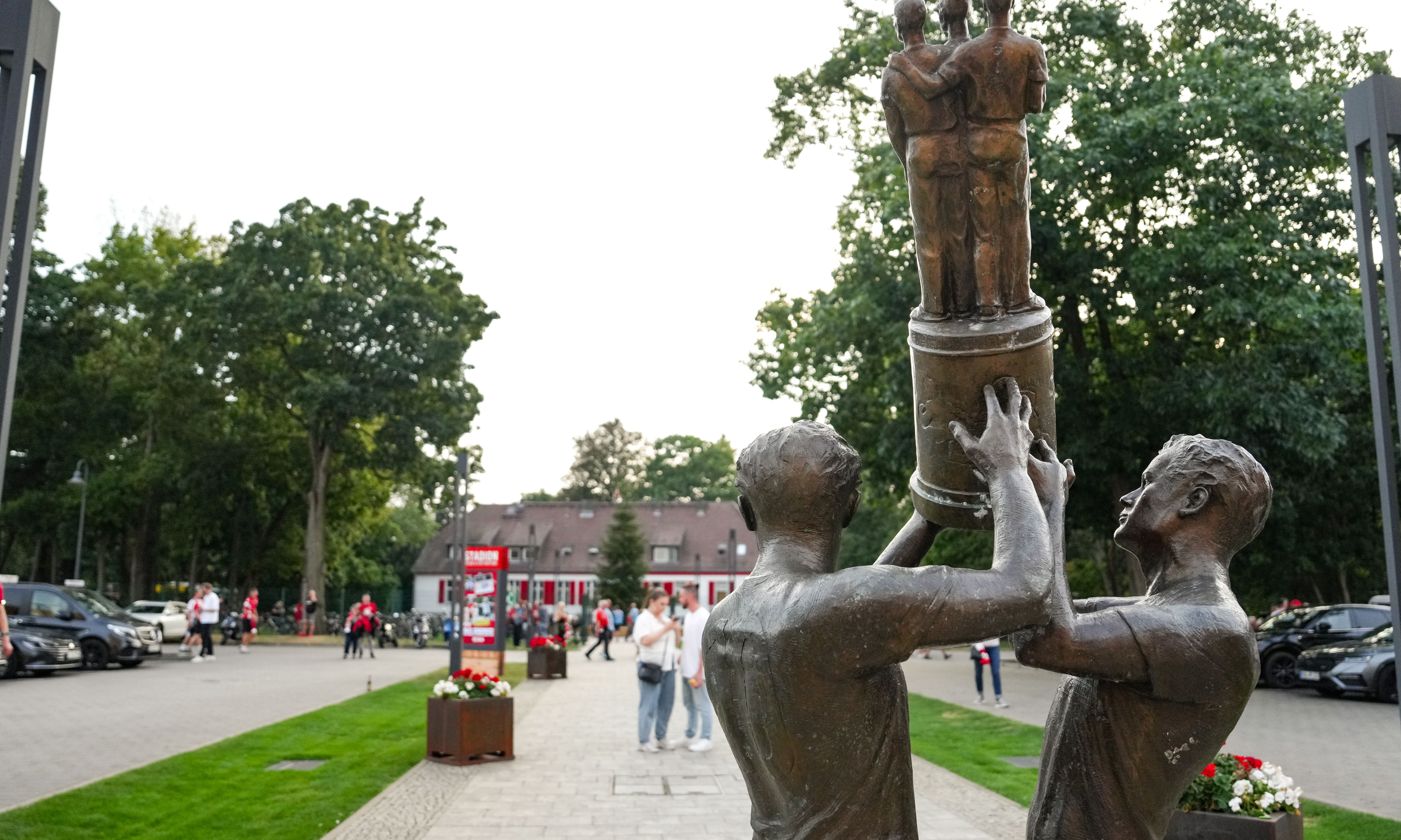 Bronzestatue von zwei Männern, die einen Pokal halten, im Hintergrund ein Park mit Bäumen und Menschen.