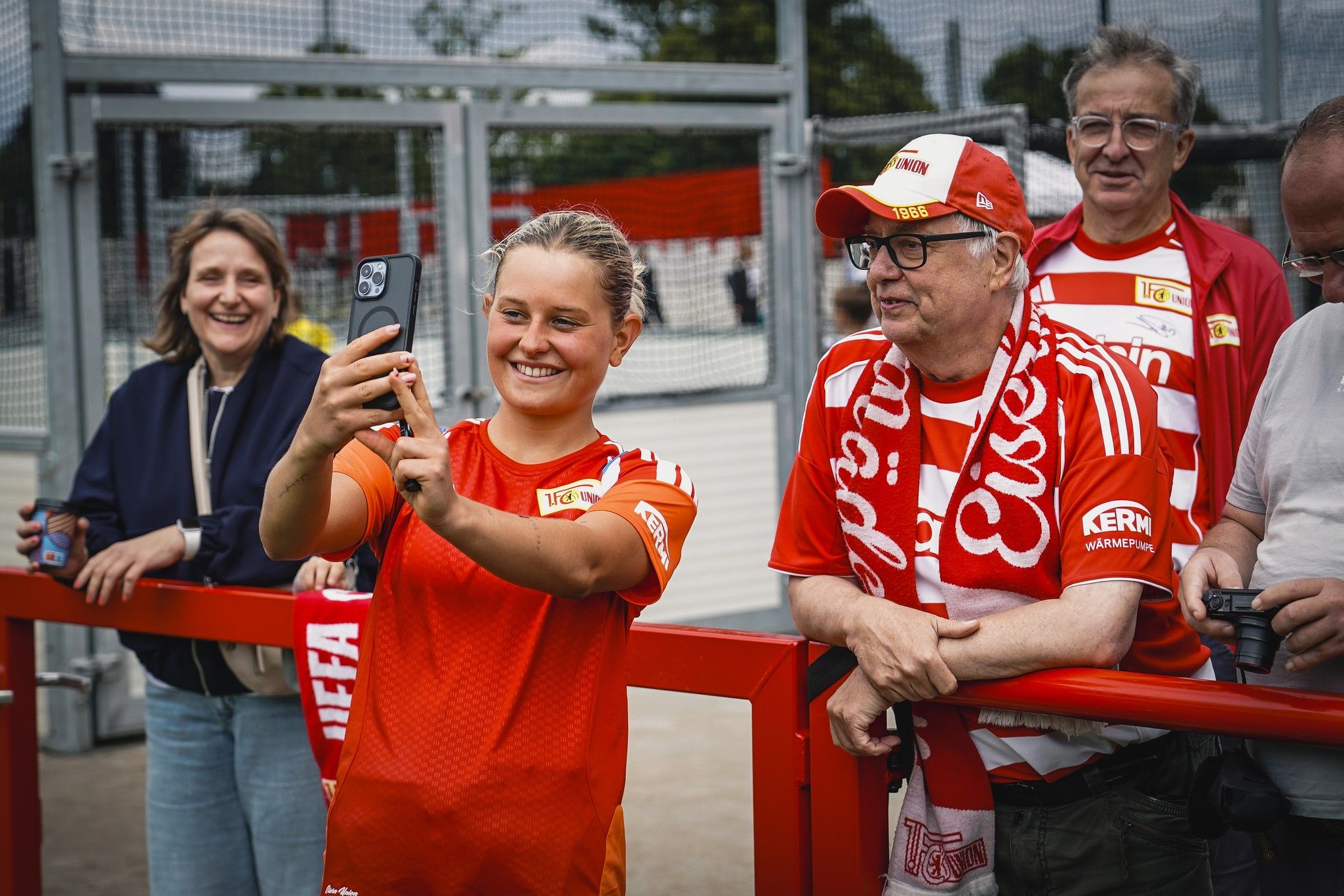 Eine lächelnde Frau in einem Fußballtrikot macht ein Selfie mit Fans, die im Hintergrund stehen und Vereinsfarben tragen.