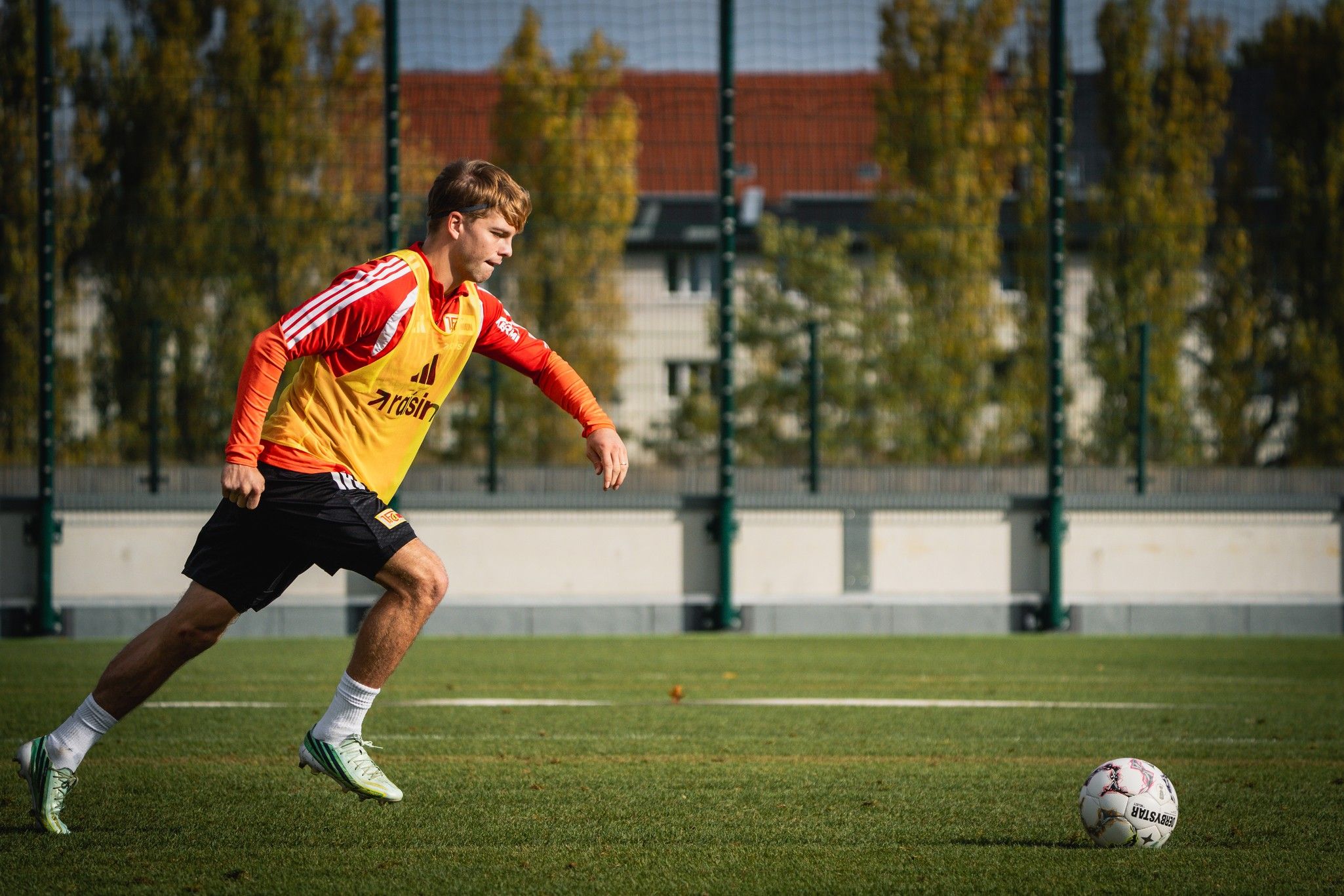 Ein junger Fußballspieler in einem orangefarbenen Trikot läuft auf einem Trainingsfeld hinter dem Ball her.